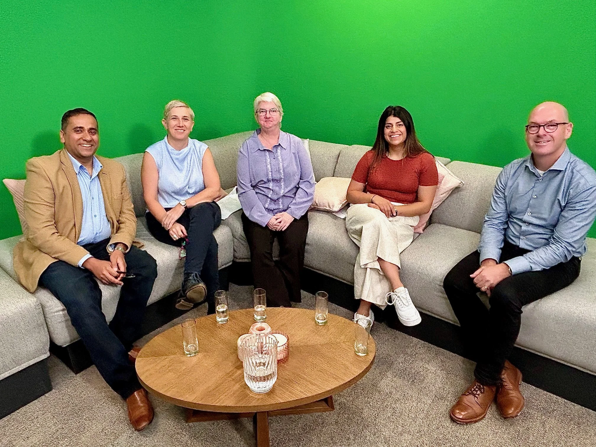 Five people sitting on a beige sectional sofa in front of a green screen, with a round wooden table in front of them holding several glasses of water and a glass pitcher.