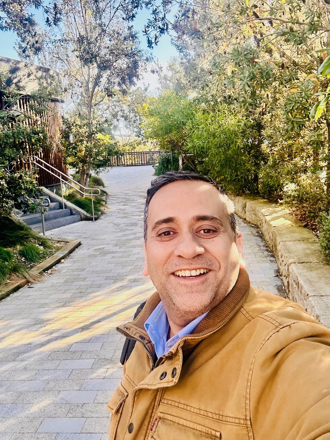 A smiling image of a man with brown jacket. In the background is a pathway with trees around it and stairs going towards a building