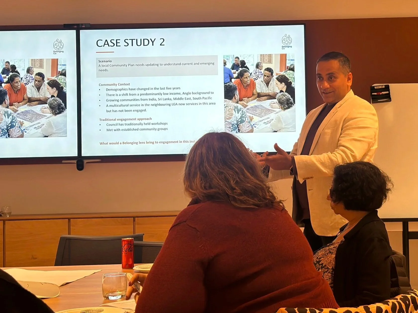 A man in a beige blazer gives a presentation to a small audience in a conference room. He stands next to a large screen displaying a slide titled "Case Study 2" that discusses community engagement and multicultural service. Several people are seated at a table, listening and taking notes, with some drinks and papers visible on the table.