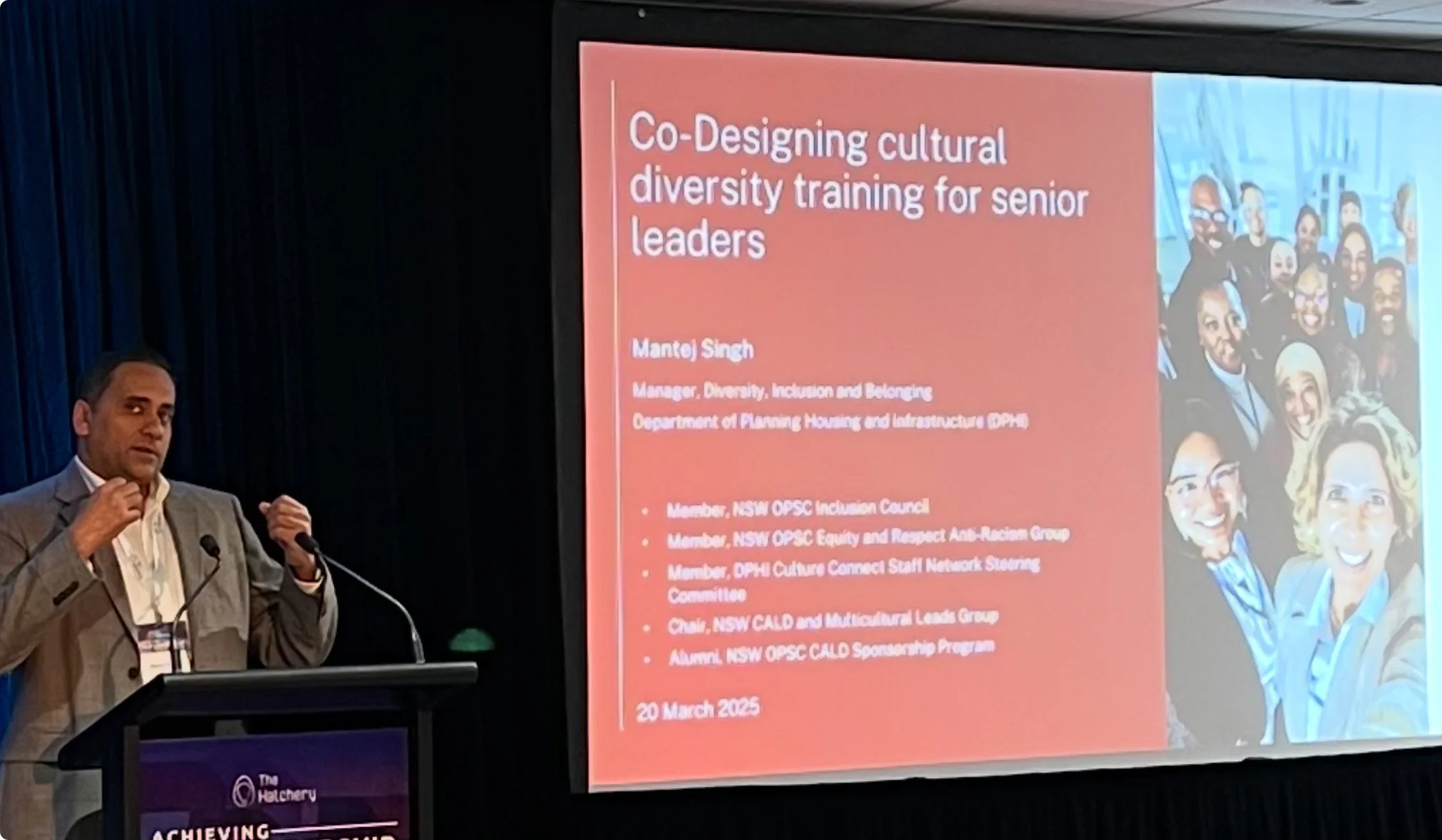 A man in a gray blazer giving a presentation at a conference, with a screen displaying a slide titled 'Co-Designing cultural diversity training for senior leaders' and a group photo of diverse people on the right side of the slide.
