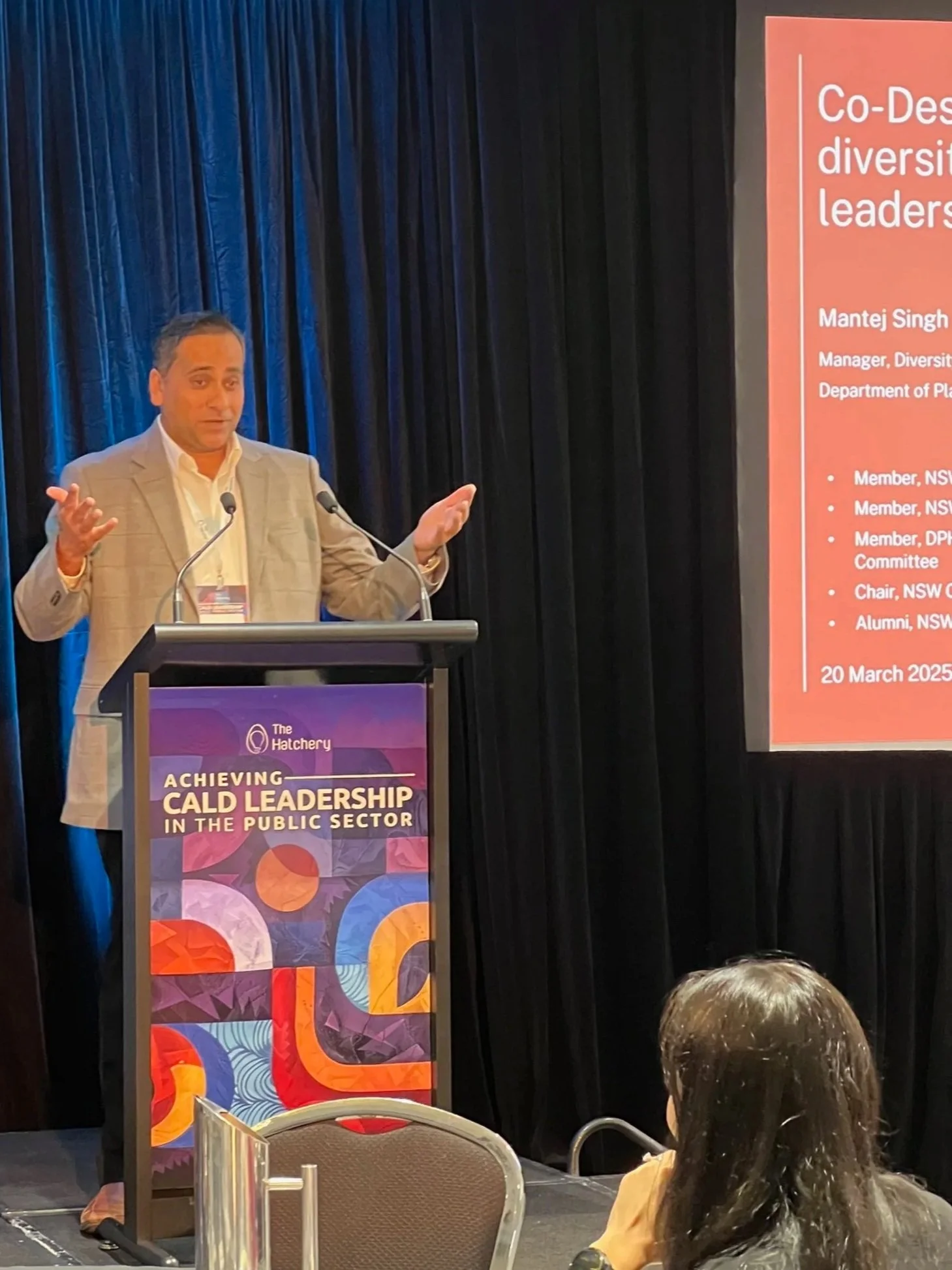 A man wearing a beige blazer and white shirt giving a speech at a conference. He is standing behind a podium with a colorful banner that reads 'Achieving CALD Leadership in the Public Sector.' Behind him are dark curtains and a large screen displaying his name, Mantej Singh, and his title, Manager, Diversity and Inclusion, Department of Planning.