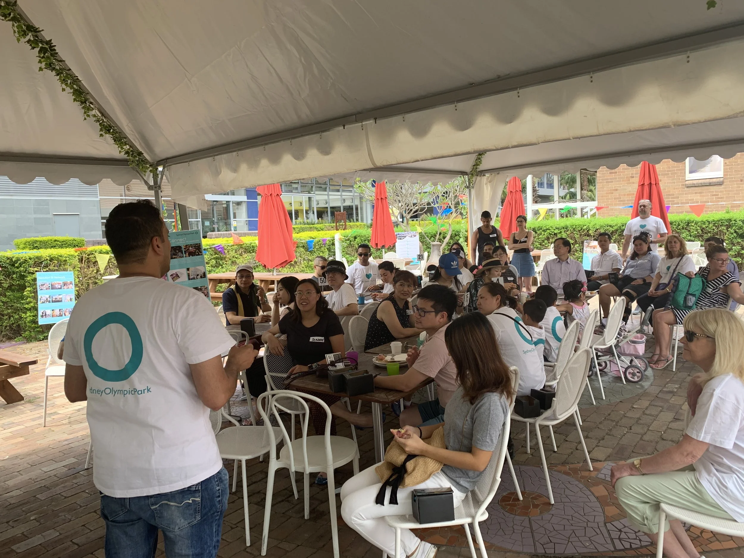 The back of person wearing white t-shirt speaking to a large group of people sitting on tables and chairs