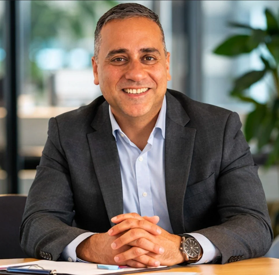 smiling image of a man with brown jacket - Belonging Co founder Mantej Singh standing outdoors with pathway and trees behind.