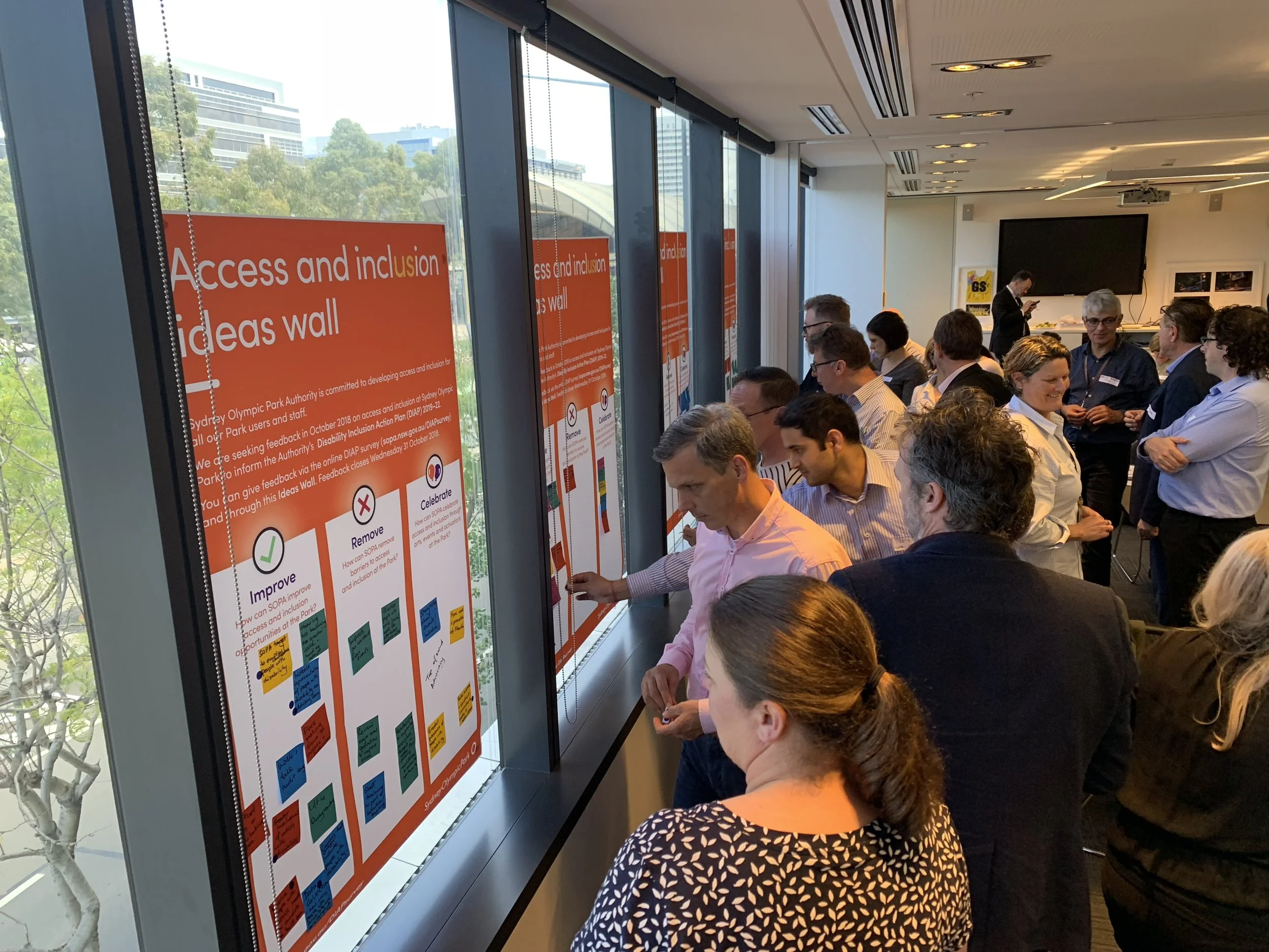 People gathered in a conference room, looking at a large attached digital display with a colorful ideas wall related to access and inclusion, with some participants engaging in discussion.