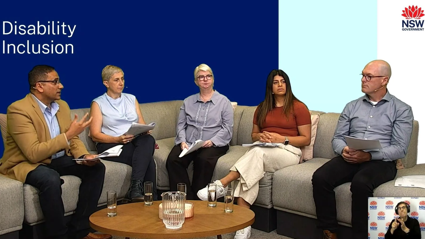 A panel discussion on disability inclusion with five diverse individuals seated on a beige couch, holding notes. Behind them is a blue and light blue backdrop with the words 'Disability Inclusion' and a logo for the NSW Government. A small inset shows a woman using sign language.