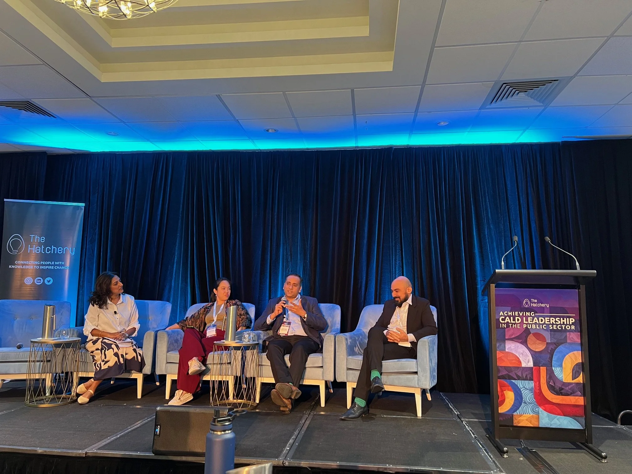 Four people seated on stage participating in a panel discussion at a conference called 'The Hatchery', with a banner and a podium displaying 'Achieving CALD Leadership in the Public Sector'. The panelists include two women and two men, one of whom is speaking into a microphone. There are small tables with water bottles and glasses in front of each panelist. The stage backdrop is black curtains with blue lighting.
