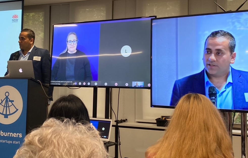 A man in a gray suit and white shirt stands at a podium with a microphone, presenting at a conference. Behind him, warped on a large screen, a woman with glasses in dark clothing is participating remotely via video call. To the right, another large screen displays a man in a blue blazer speaking into a microphone. The audience is seated facing the stage.