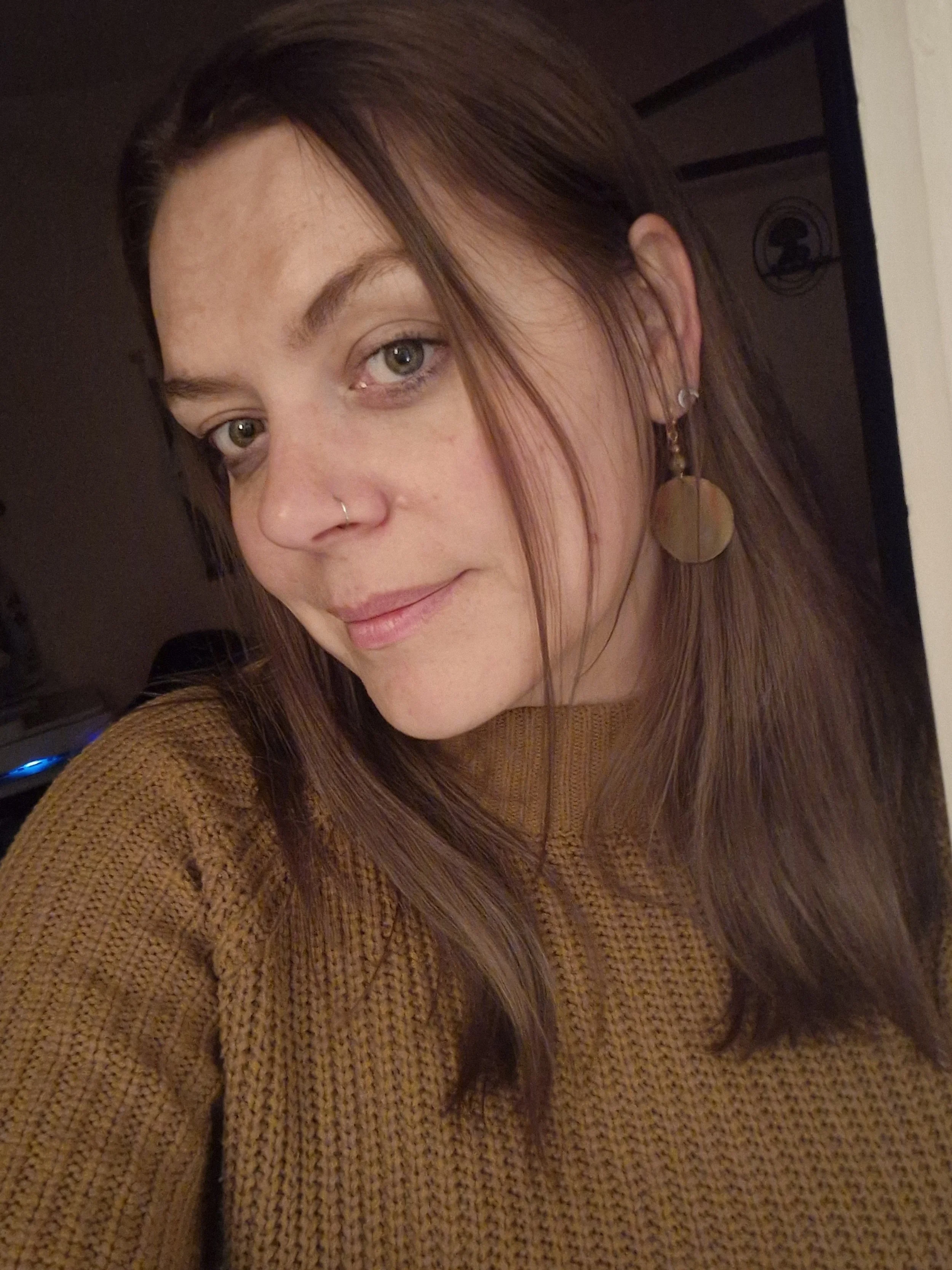 A woman with shoulder-length brown hair and light-colored eyes, wearing a brown sweater, silver earrings, and a small nose ring, smiling slightly at the camera.