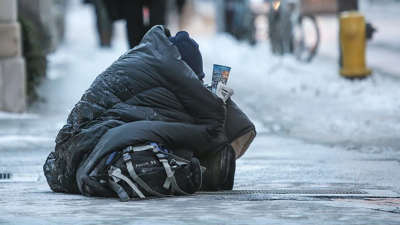 A homeless person sits on a city sidewalk in winter, wrapped in a dark sleeping bag, holding a cup, with a backpack nearby.