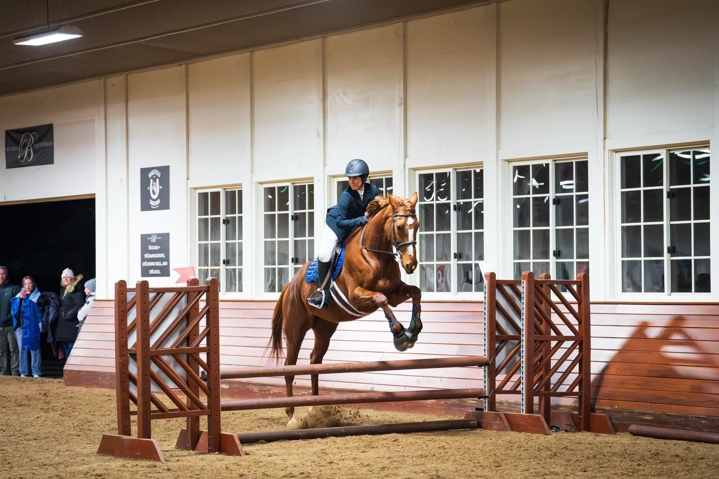 A rider wearing a helmet and riding gear guides a bay horse over a jump inside an indoor equestrian arena. Spectators stand nearby watching.