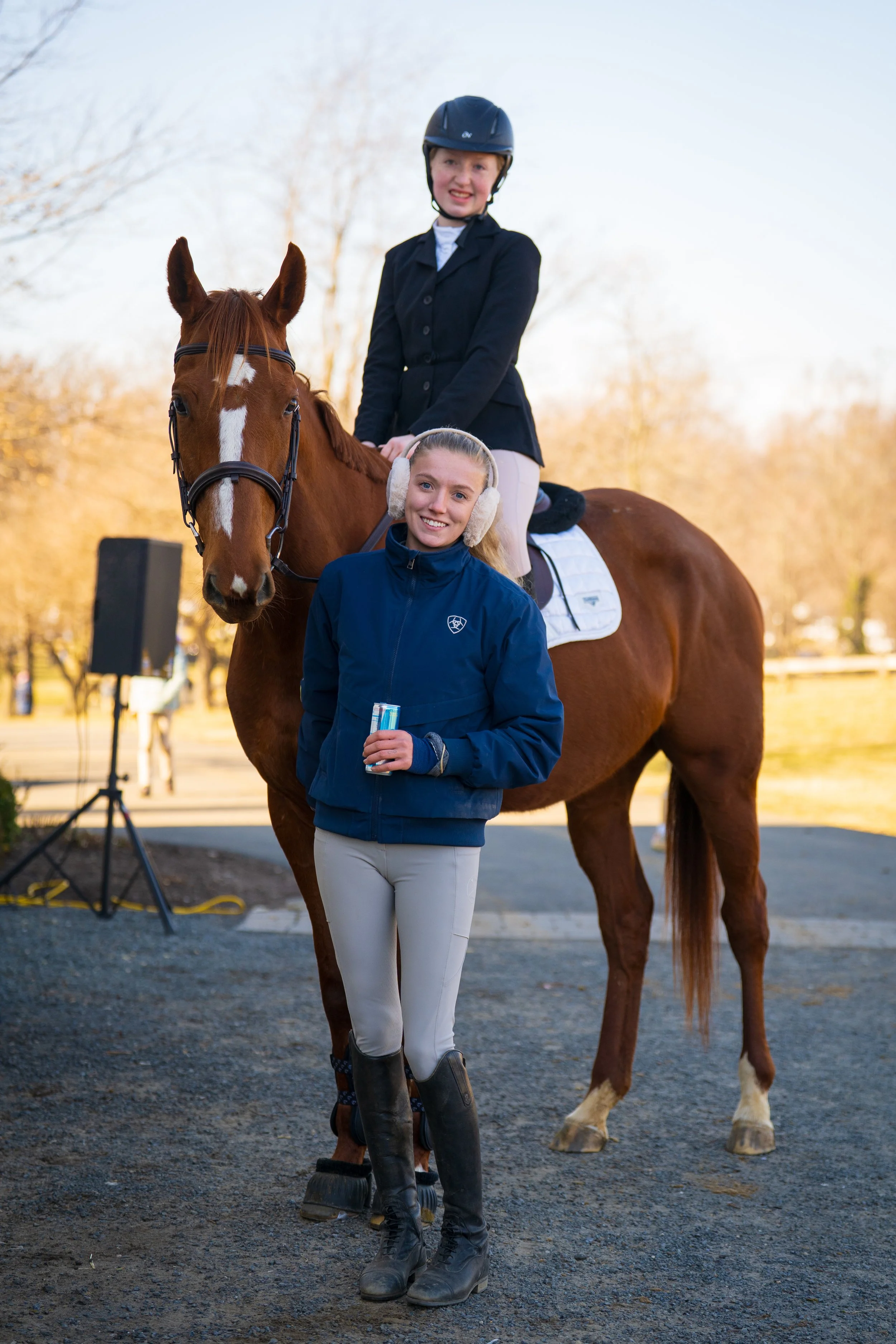 Two young women at an outdoor horse riding event, one seated on a chestnut horse wearing a black helmet and riding jacket, the other standing in front wearing ear protection, a blue jacket, holding a drink, with trees in the background.