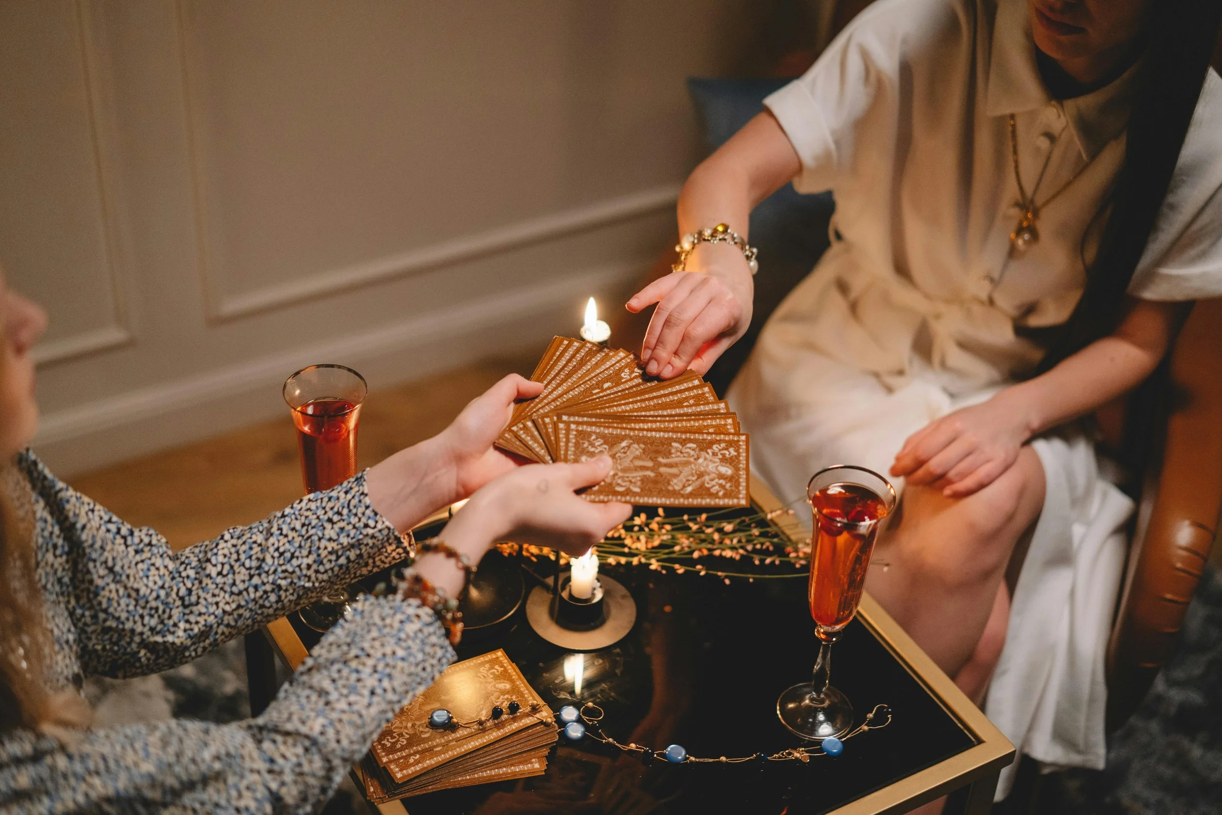 Two women sitting at a coffee table with tarot cards, candles, and drinks, engaging in a tarot reading or card game.