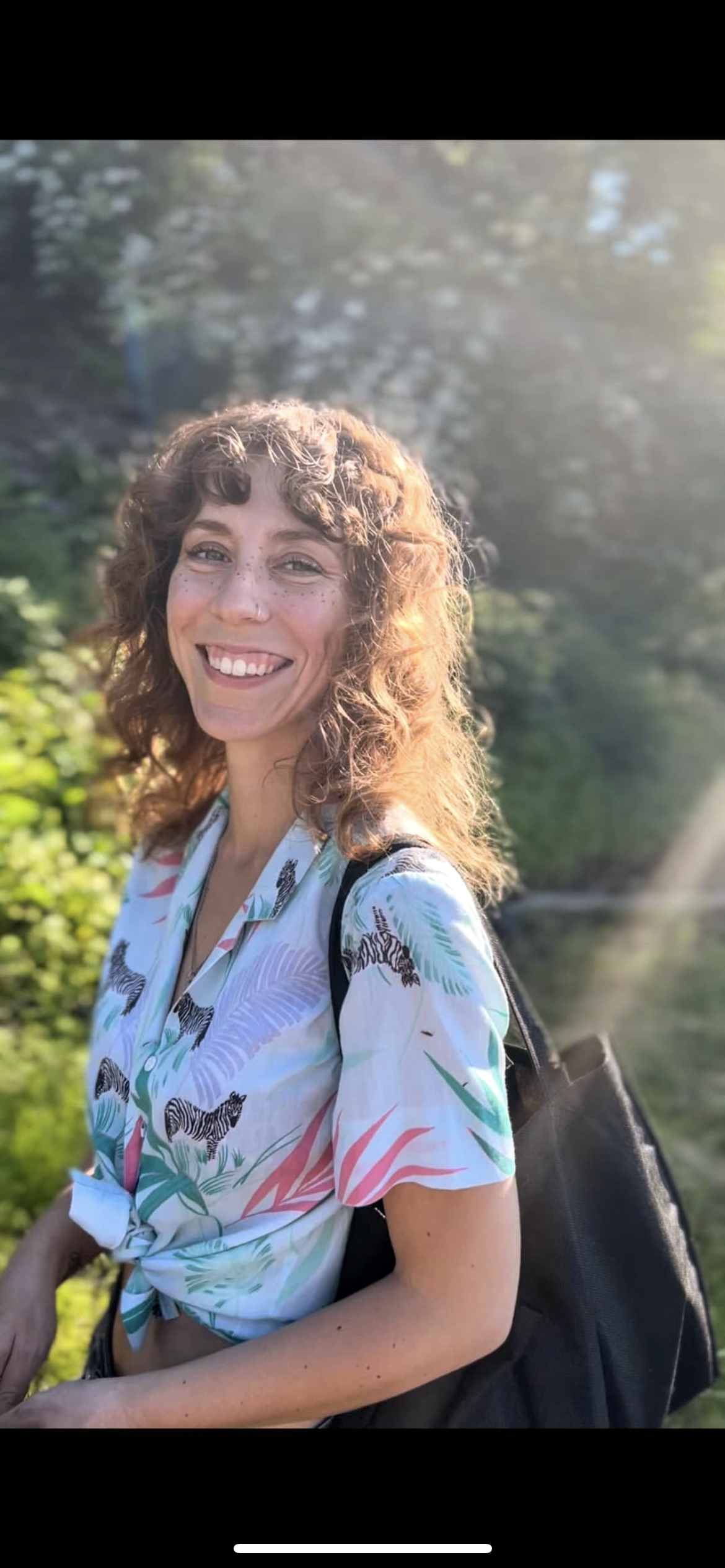 A smiling woman with curly brown hair and freckles, wearing a colorful shirt with zebras and jungle patterns, standing outdoors with greenery and sunlight in the background.