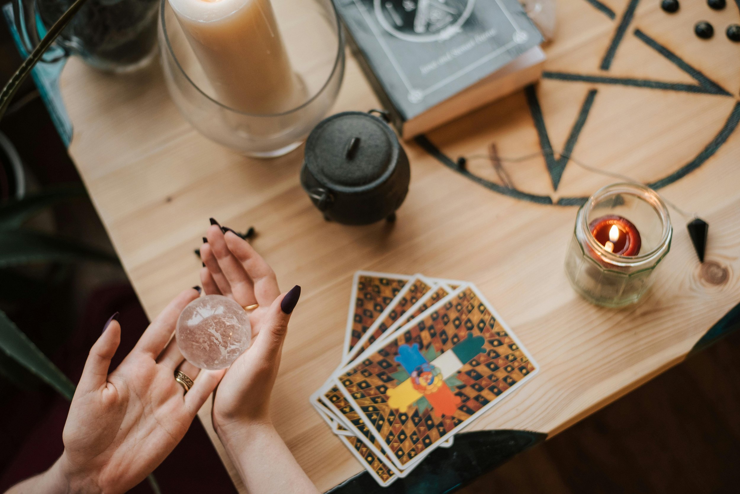 A person holding a crystal ball over a wooden table with tarot cards, candles, and various decorative items.