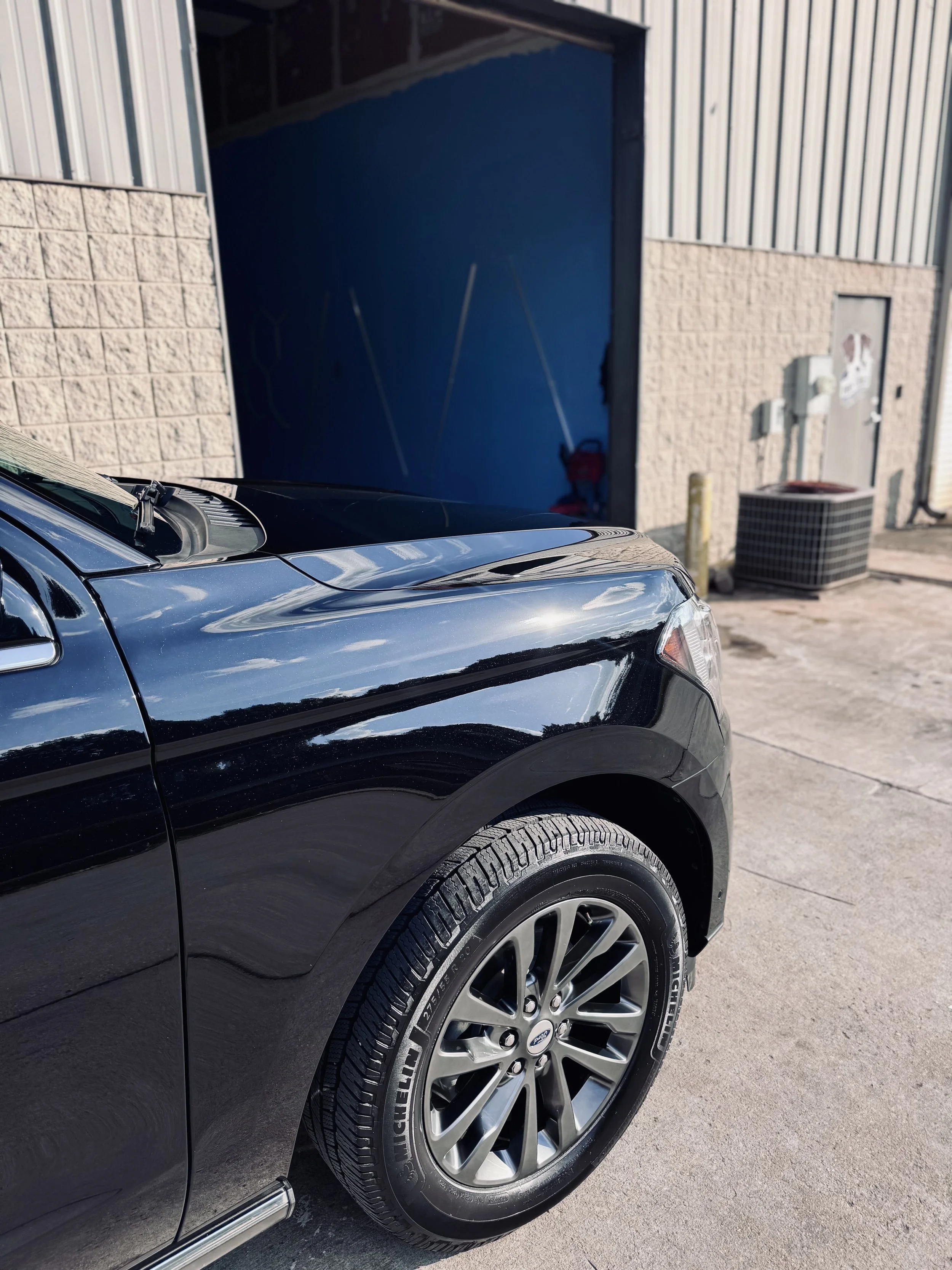 Close-up of the front section of a black car, showing the wheel and part of the hood, parked outside a building with a blue door and brick wall.