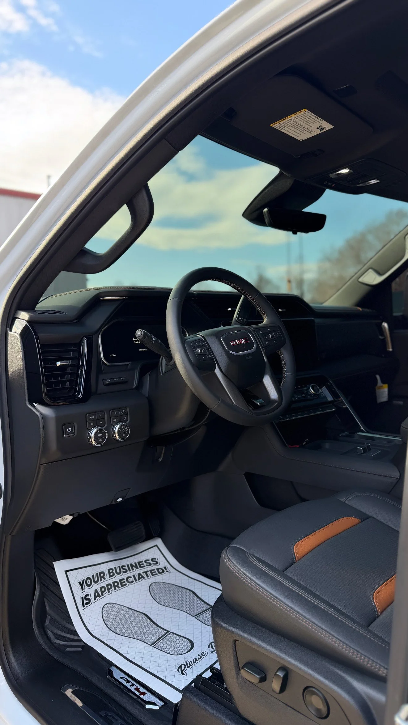 Interior view of a GMC vehicle's driver's side, showing the steering wheel, dashboard, and front seat with a paper floor mat that reads 'Your Business Is Appreciated! Please Drive Safely.' and features footprints.