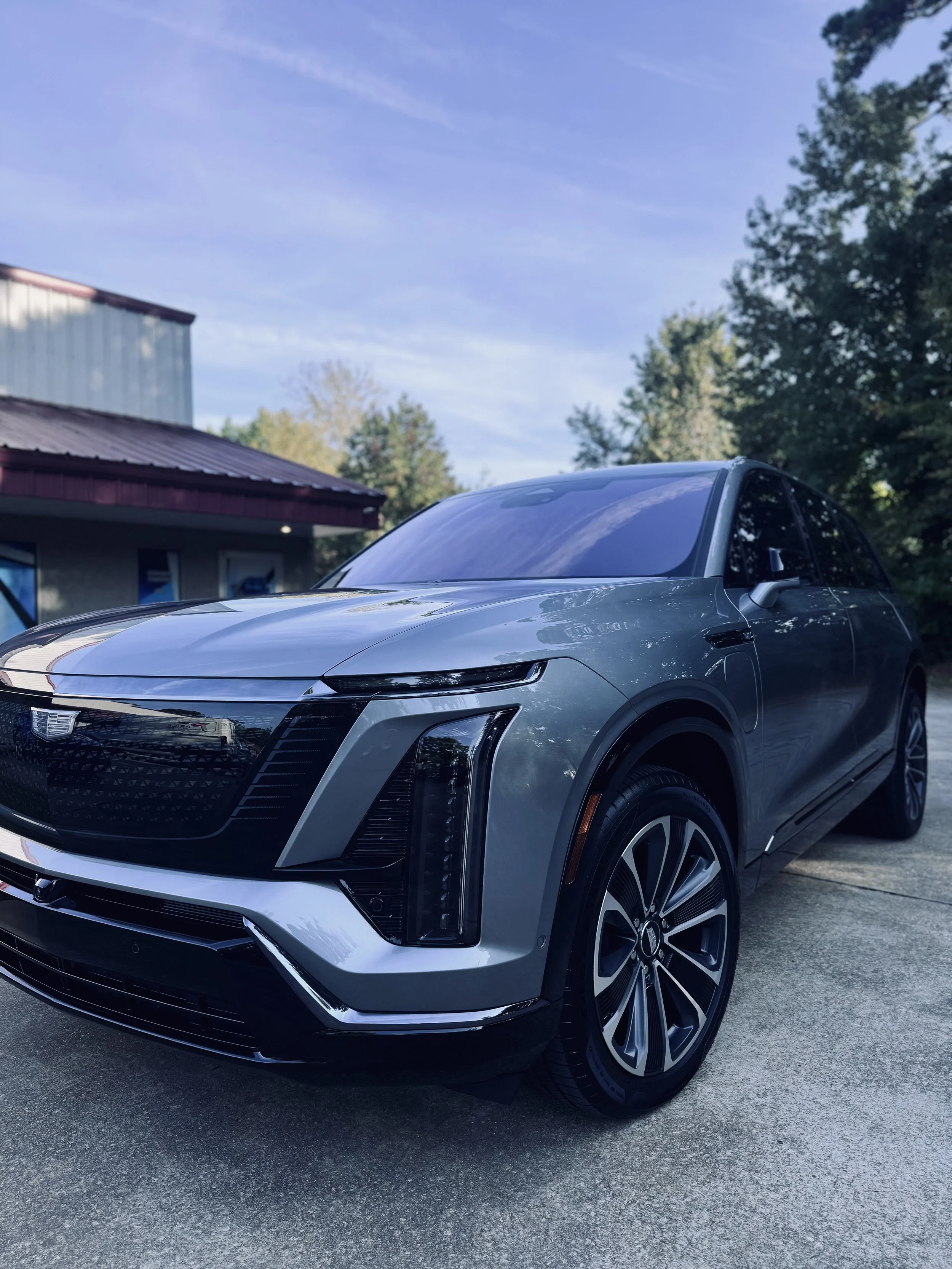 A gray electric SUV parked on a driveway with a building and trees in the background under a partly cloudy sky.