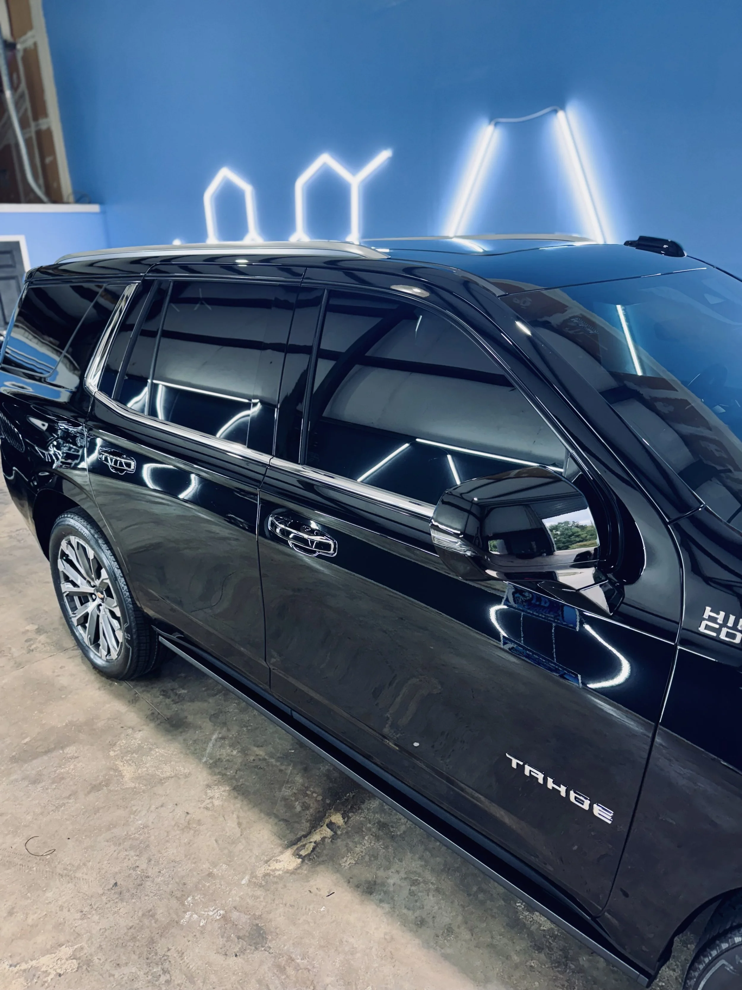Black Chevrolet Tahoe parked indoors with blue walls and neon light decorations reflected on its surface after tint