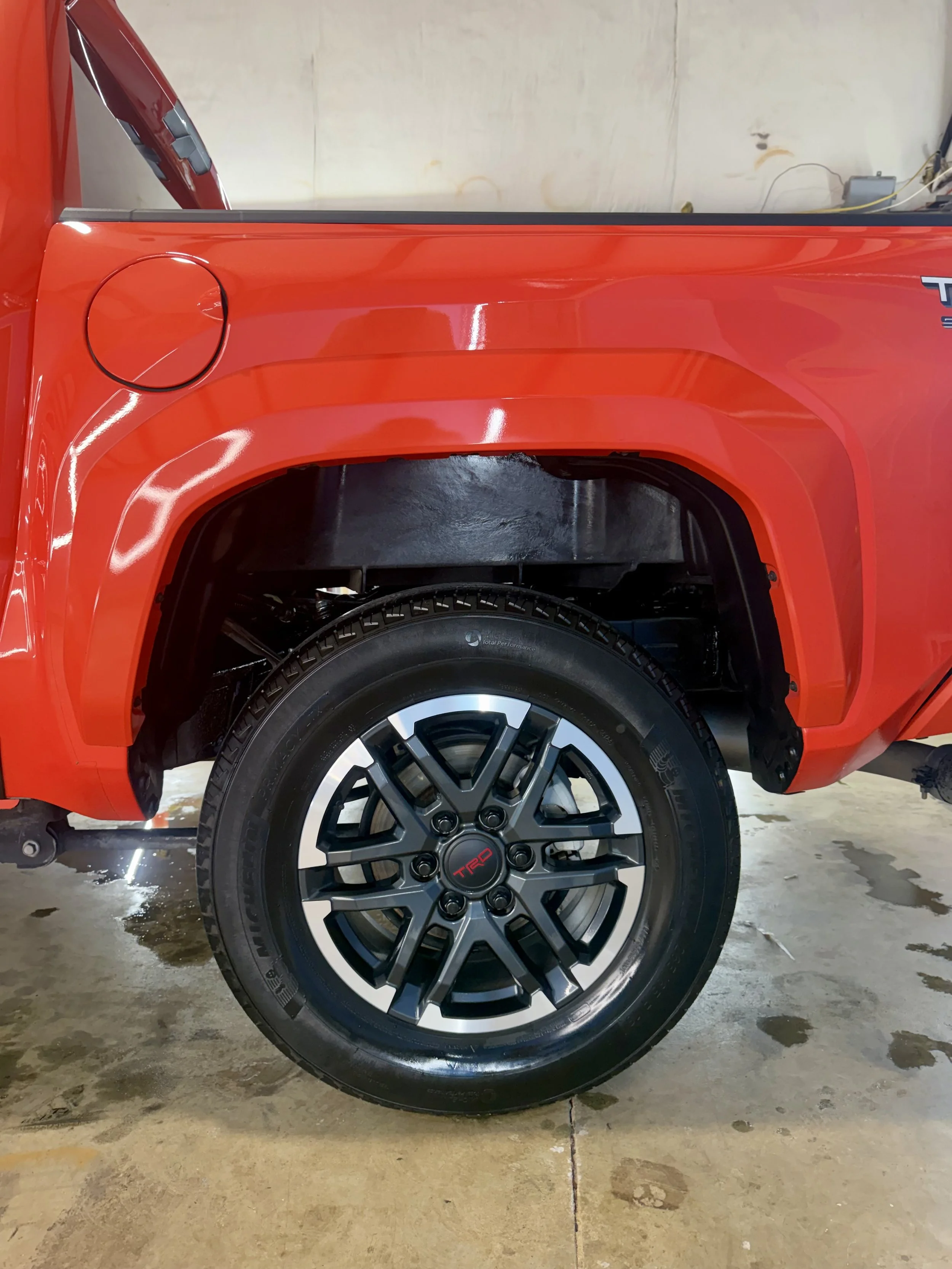 Close-up of a red vehicle's rear wheel and part of the body, with a black alloy wheel and tire, parked on concrete after detailing and vinyl
