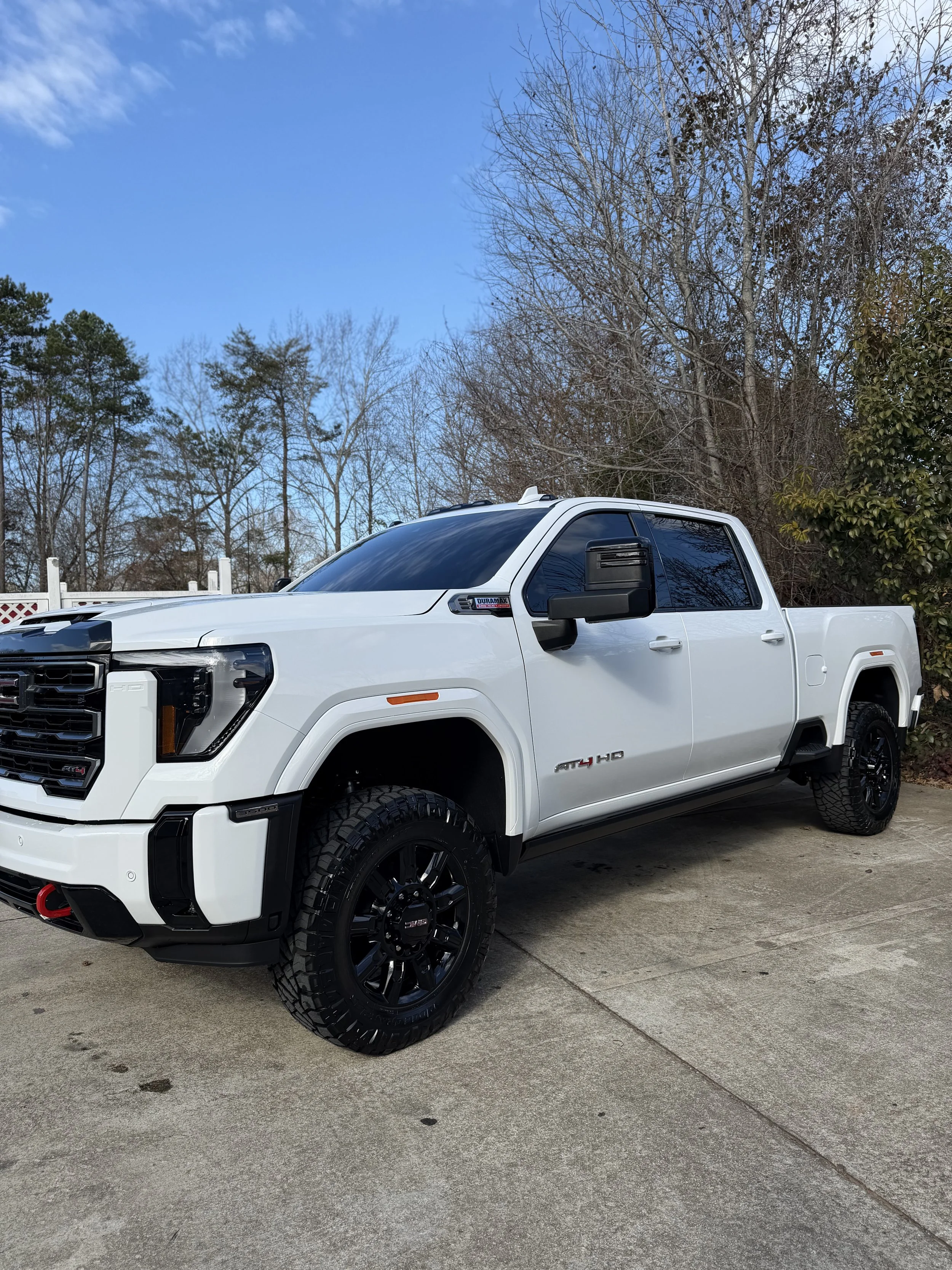 A white GMC heavy-duty pickup truck with black accents parked on a concrete driveway, with trees and a blue sky in the background.