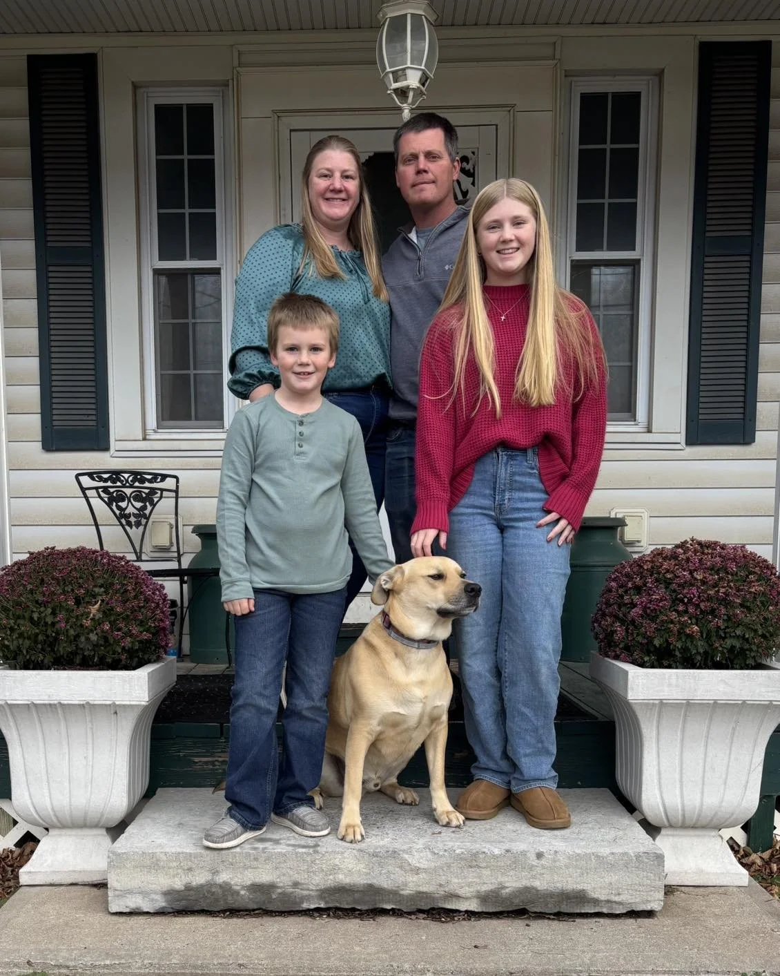 Family portrait of four people and a dog standing in front of a house porch, with potted plants on each side. The family includes two adults, one teenage girl, and one young boy.