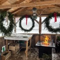 Decorated outdoor wooden pergola with Christmas wreaths and red ribbons, a small fire pit with flames, and cardboard boxes on a shelf, winter scene outside.