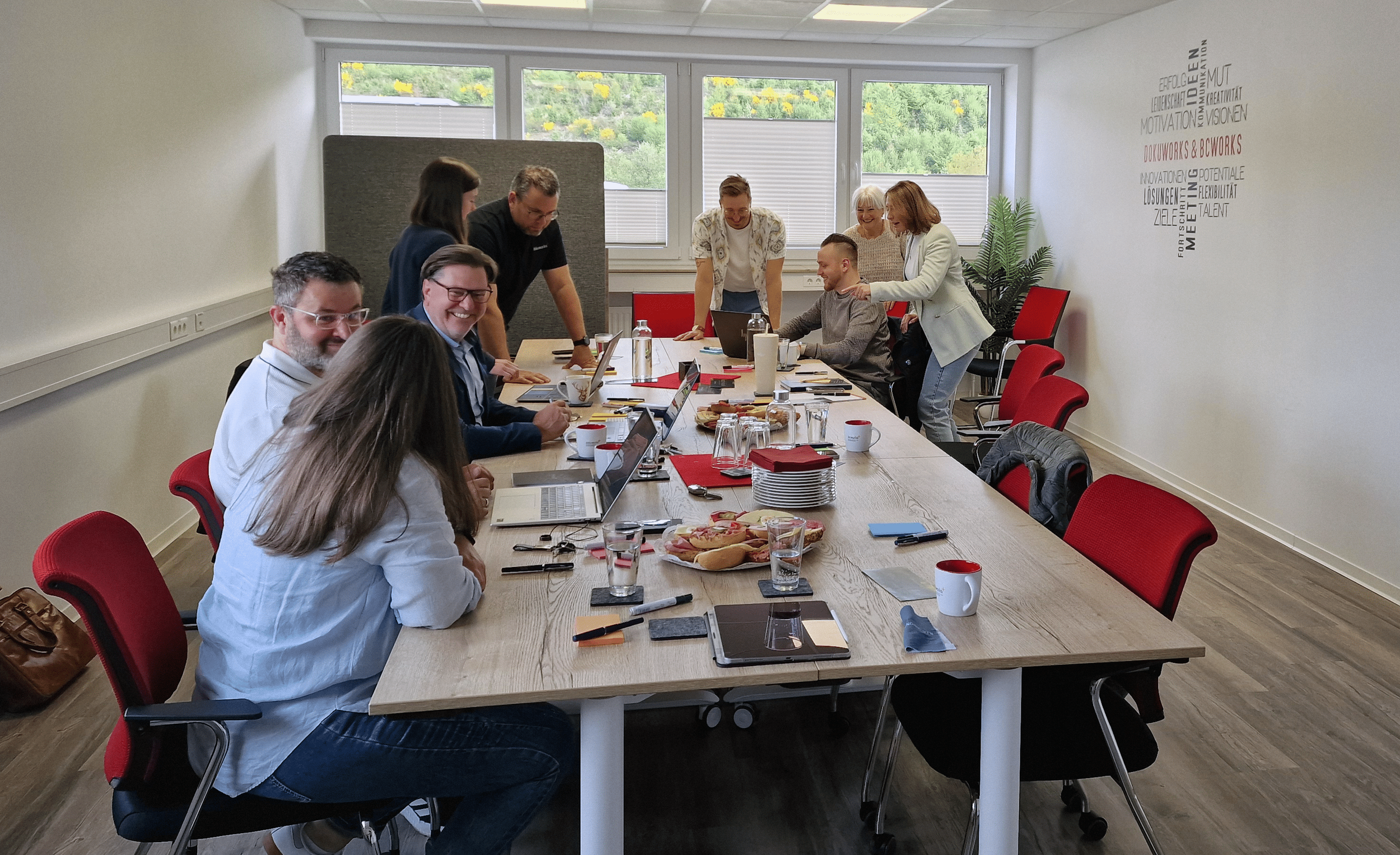 Mehrere Personen sitzen um einen langen Tisch in einem Meetingraum, während einige andere stehen. Es gibt Laptops, Notizblöcke, Tassen, Wasserflaschen und Snacks auf dem Tisch. Die Stimmung ist fröhlich und kollaborativ.