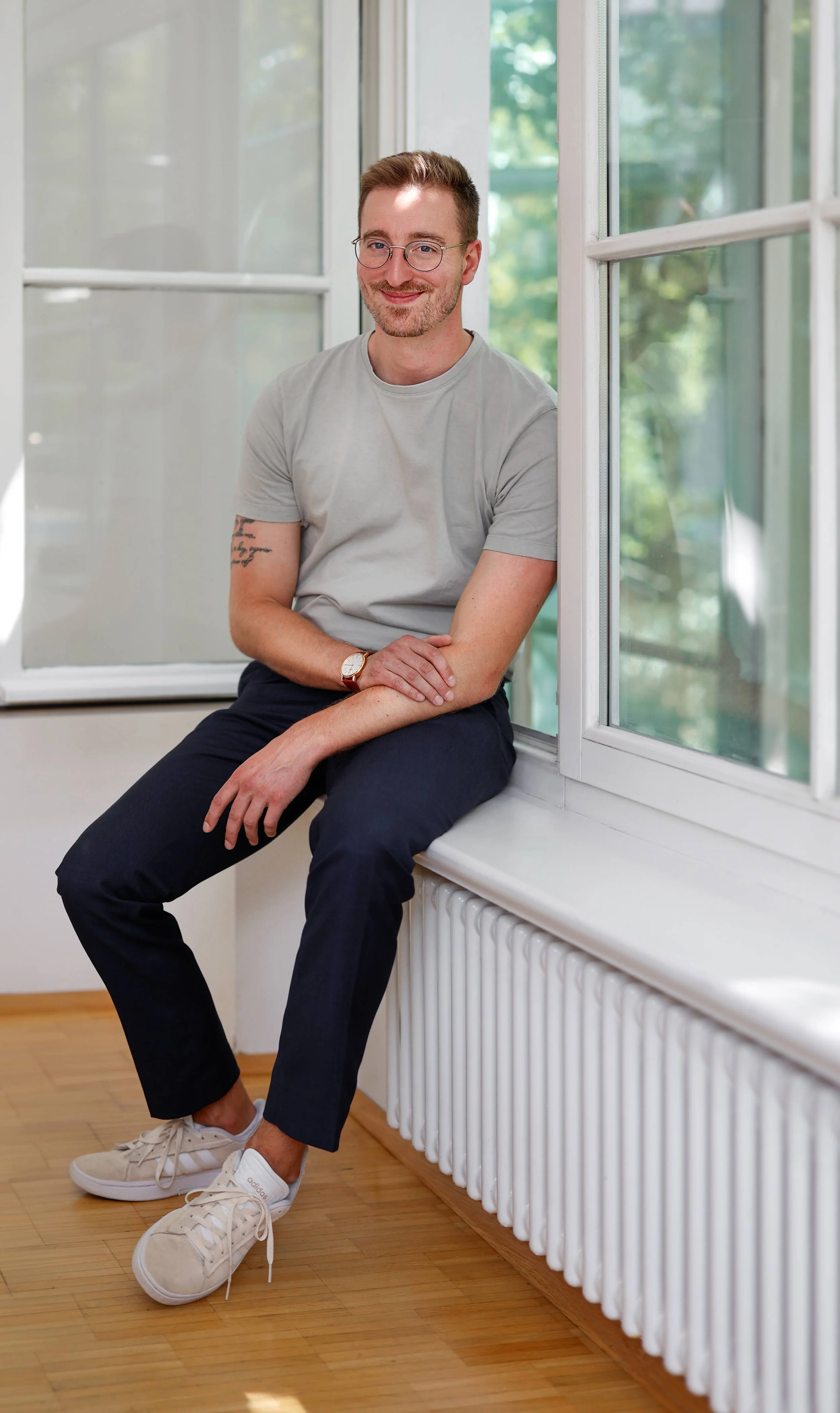 Inhaber Björn Müsse sitzend auf einer Fensterbank, lächelnd, casual Kleidung, Brille, Armbanduhr, Tattoos, helle Haare, Holzfußboden, große Fenster mit Blick nach draußen, grüne Blätter im Hintergrund.