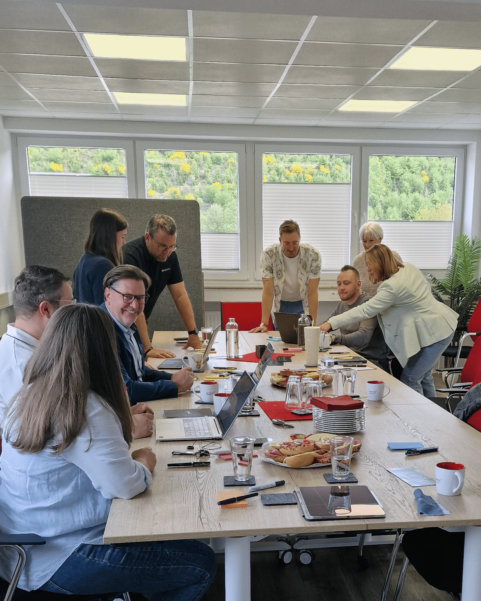 Menschen sitzen um einen langen Tisch in einem Büro, diskutieren und arbeiten gemeinsam. Auf dem Tisch befinden sich Laptops, Wasserflaschen, Tassen und Snacks.