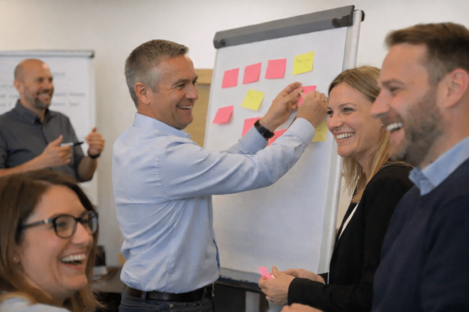 Menschen in einem Meetingraum, die sich an einem Whiteboard mit bunten Post-its beteiligen und lachen.