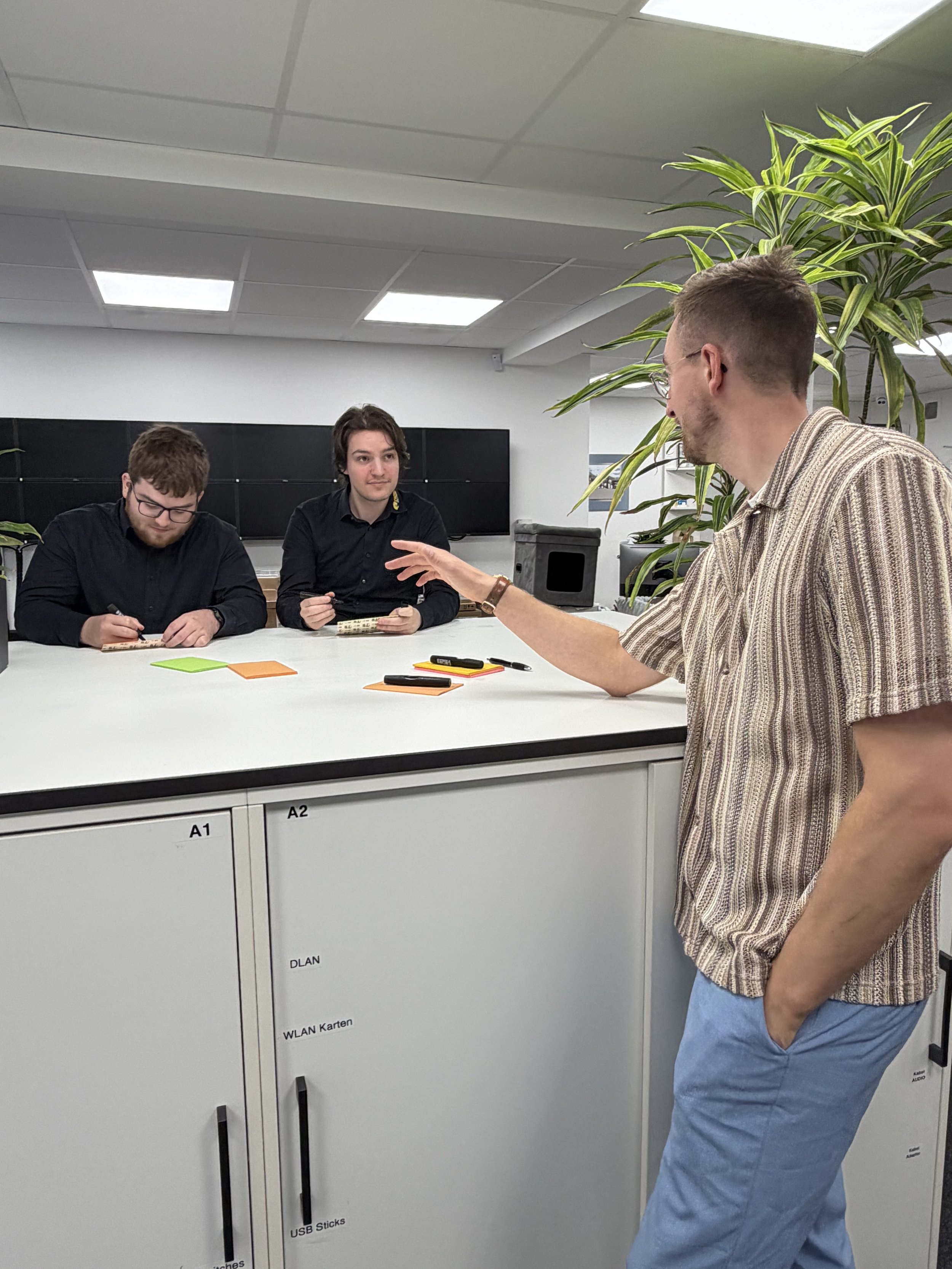 Drei junge Männer sitzen an einem weißen Tisch und führen ein Gespräch. Ein weiterer Mann steht am Tisch, seine Hand gestikuliert während er spricht. Im Hintergrund sind Pflanzen und eine schwarze Tafel sichtbar.