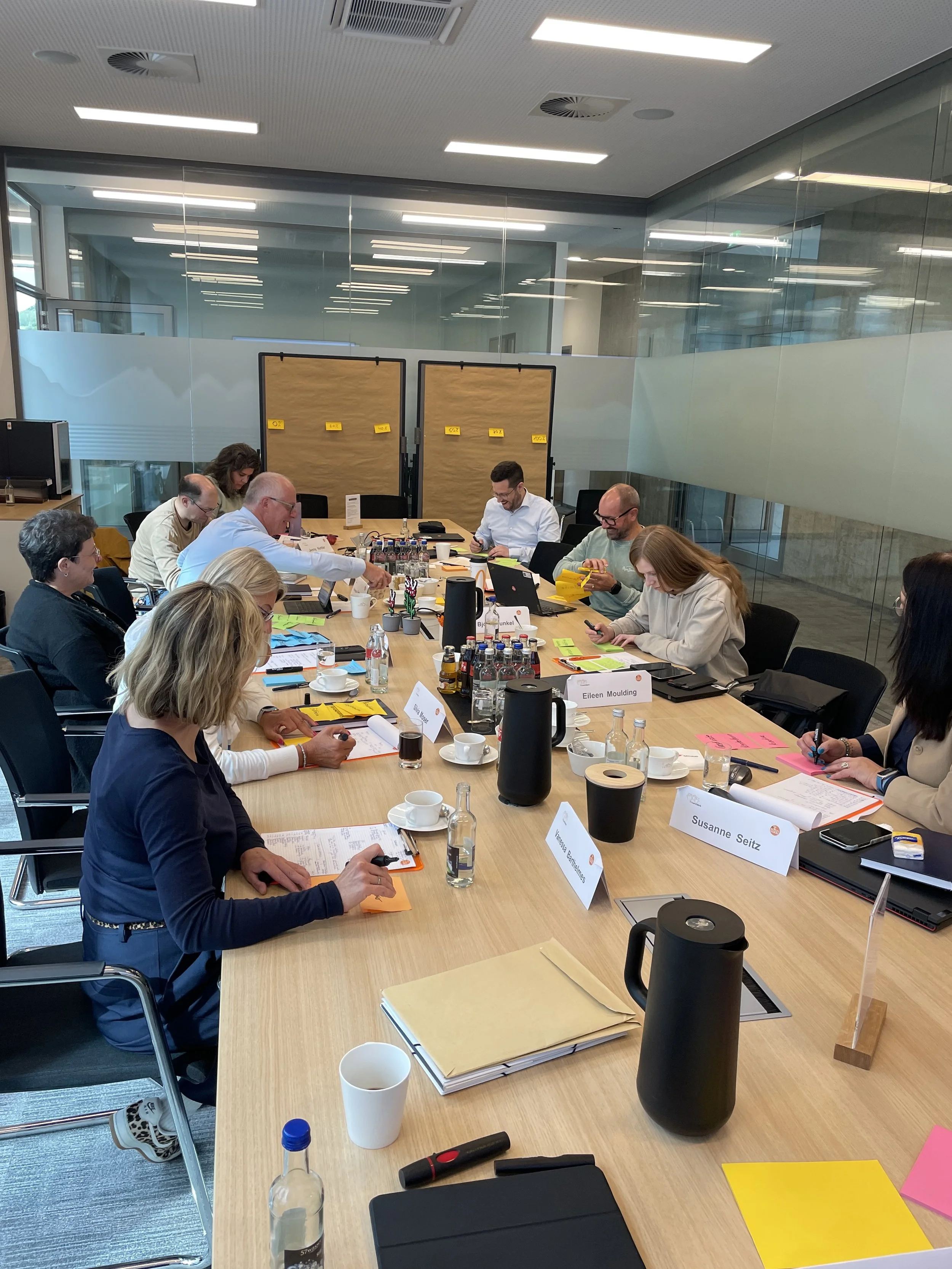 Mehrere Personen sitzen um einen Konferenztisch bei einem Meeting. Der Tisch ist mit Getränken, Notizmaterialien, Laptops und anderen Gegenständen bedeckt. Im Hintergrund sind Namensschilder und Pinnwände zu sehen.