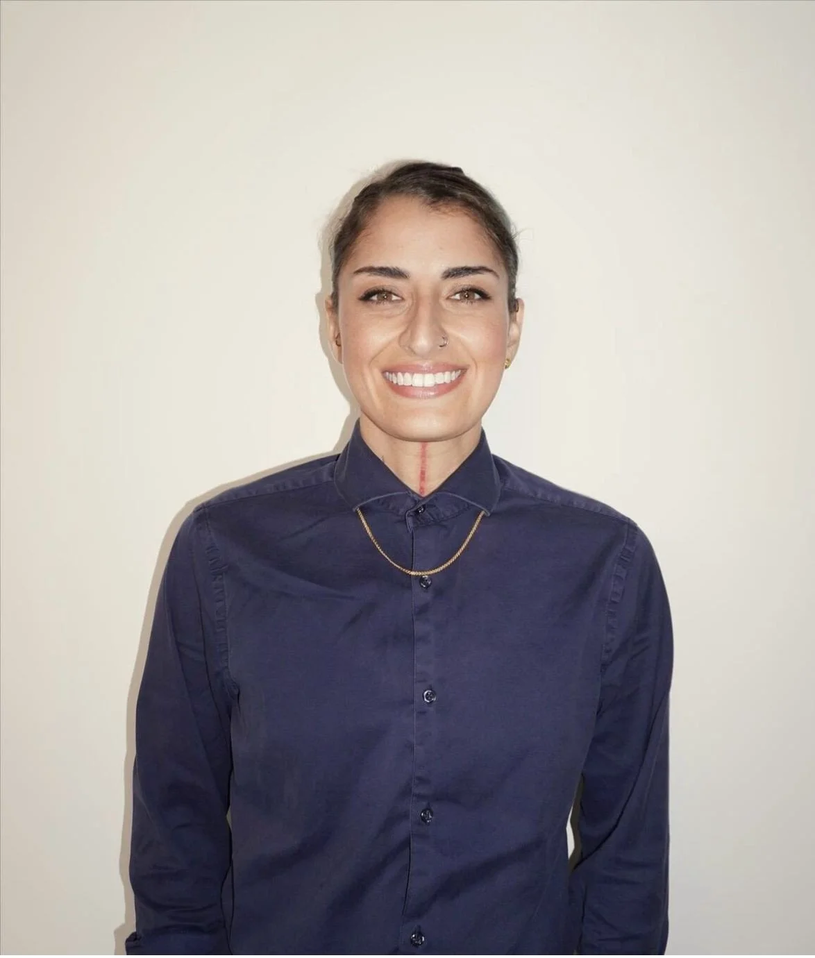 Portrait of a smiling woman with dark hair tied back, wearing a navy blue button-up shirt, gold chain, and small earrings, standing against a plain light-colored background.