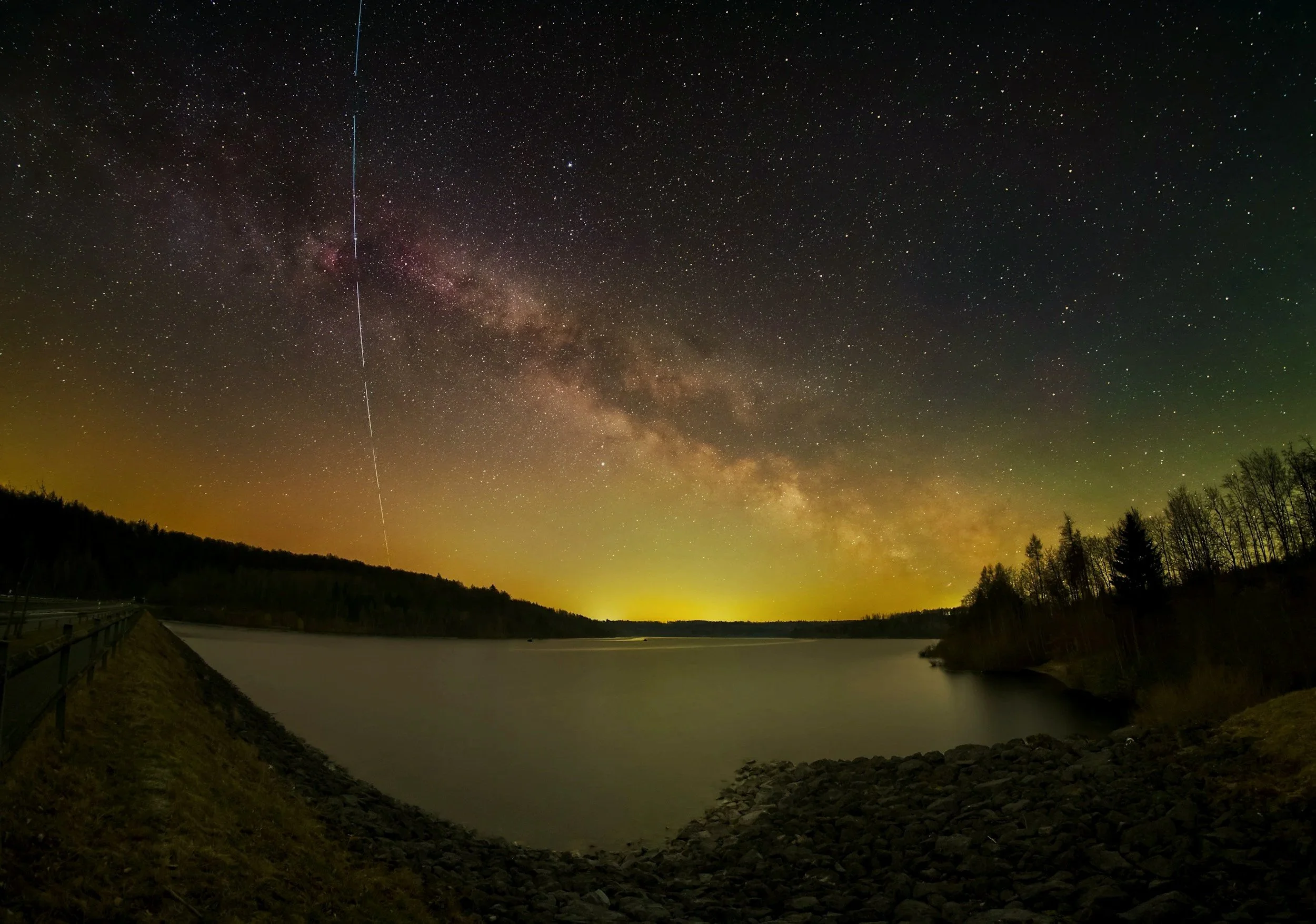 Night sky over a lake with the Milky Way galaxy, stars, and a meteor streaking across the sky. Hills and trees are silhouetted along the shoreline.