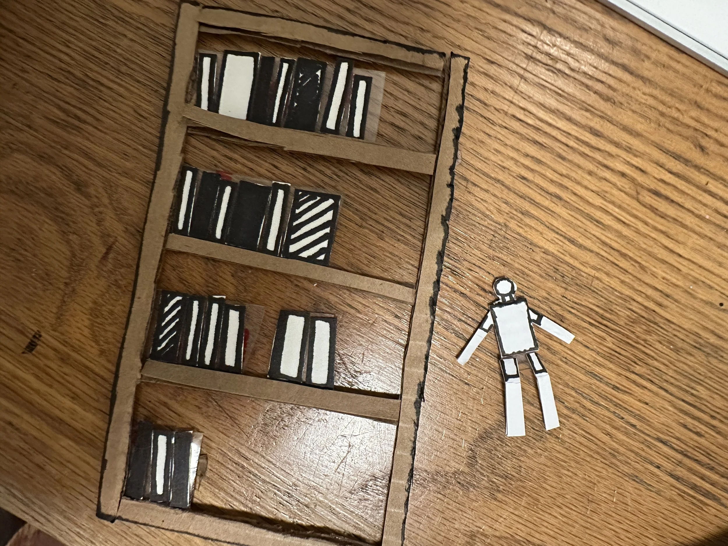 Handmade paper craft depicting a bookshelf with black and white books and a paper figure lying on a wooden floor.