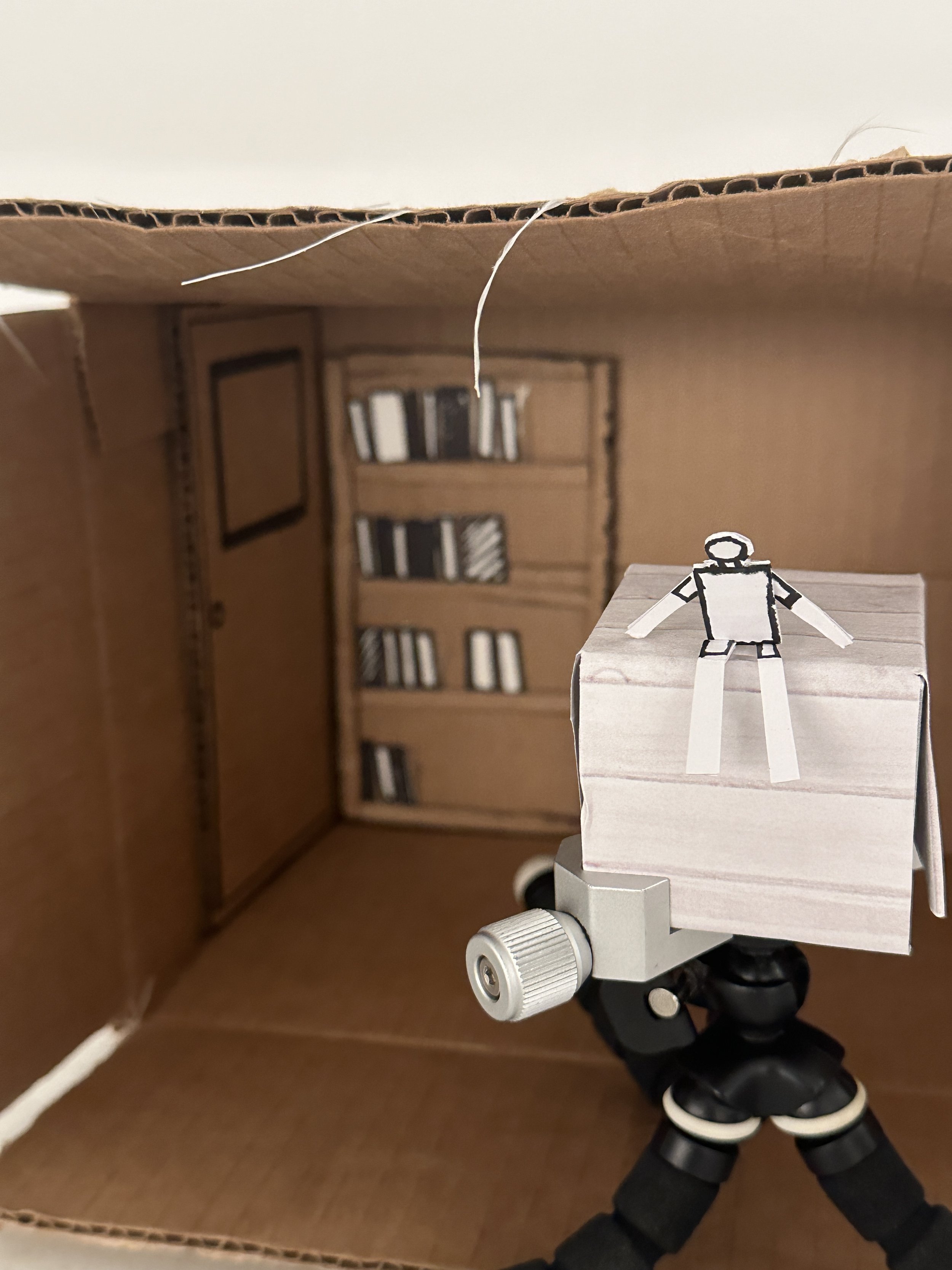 A miniature cardboard room with a bookshelf and a wall picture, seen from inside a protractor tripod with a paper figure on top.