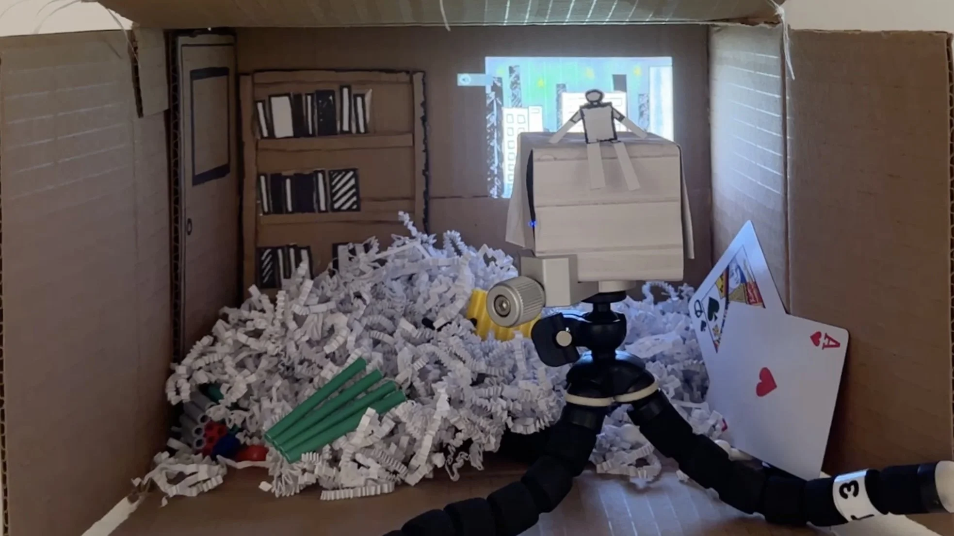 A diorama of a room built inside a cardboard box, featuring a small camera on a tripod, some playing cards, a pile of white shredded paper, and a drawn bookshelf with books on the back wall.