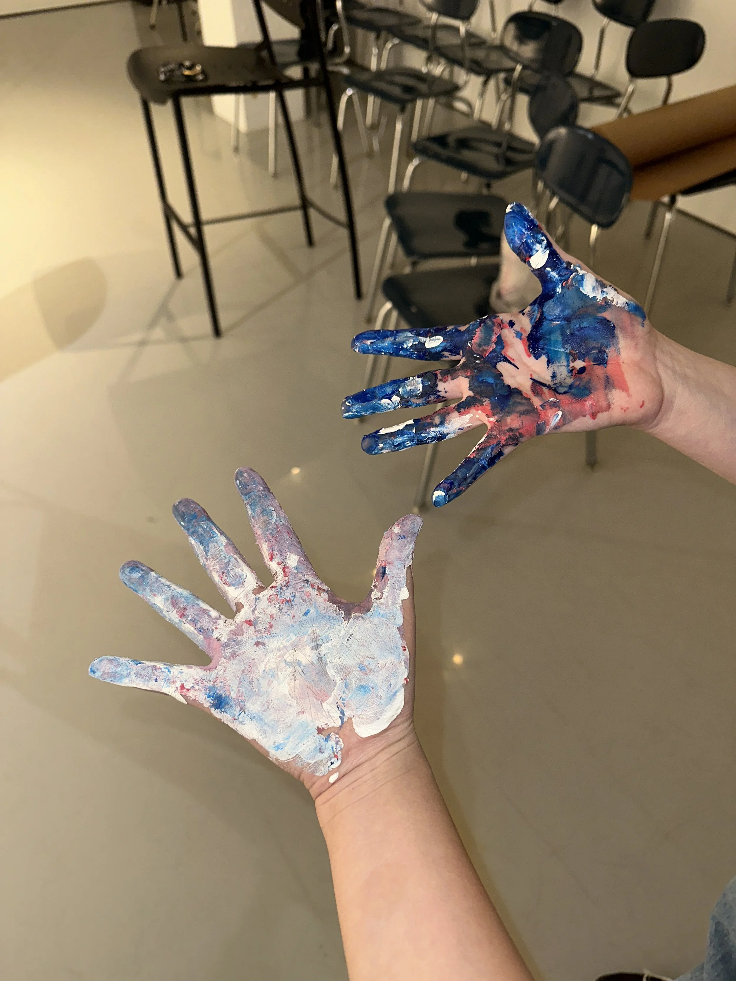 Two hands covered in colorful paint, with one hand showing mostly white and pink paint and the other with blue and pink paint, in an indoor room with black chairs and a table in the background.