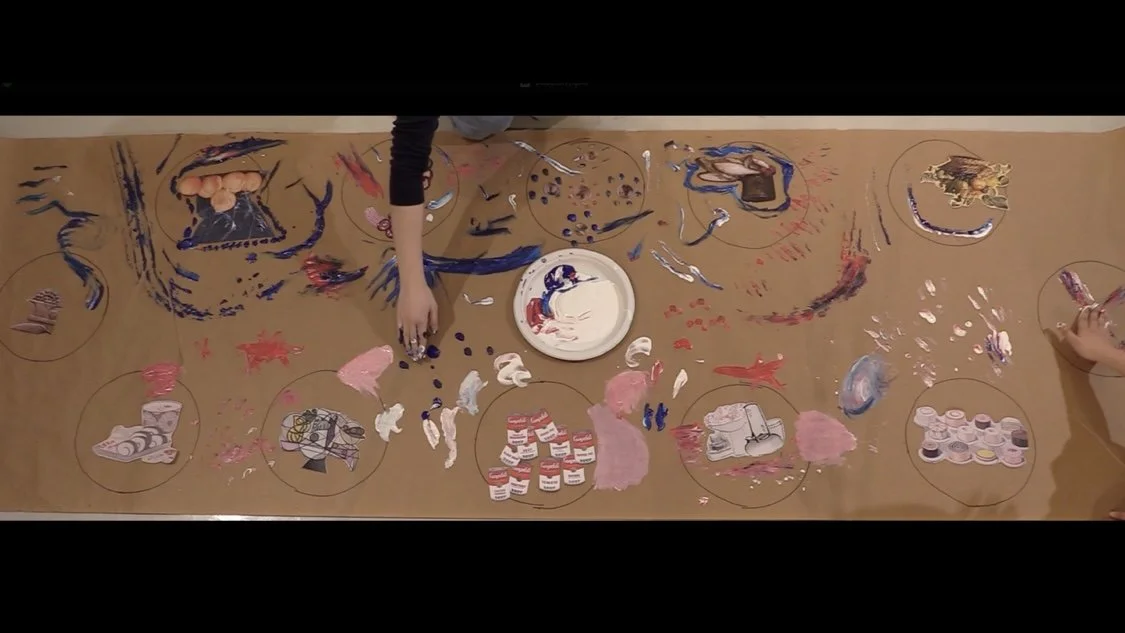 People are painting and decorating a long brown table with various art supplies, including paints, brushes, and paper cutouts. The table is covered with colorful paint and artistic designs.