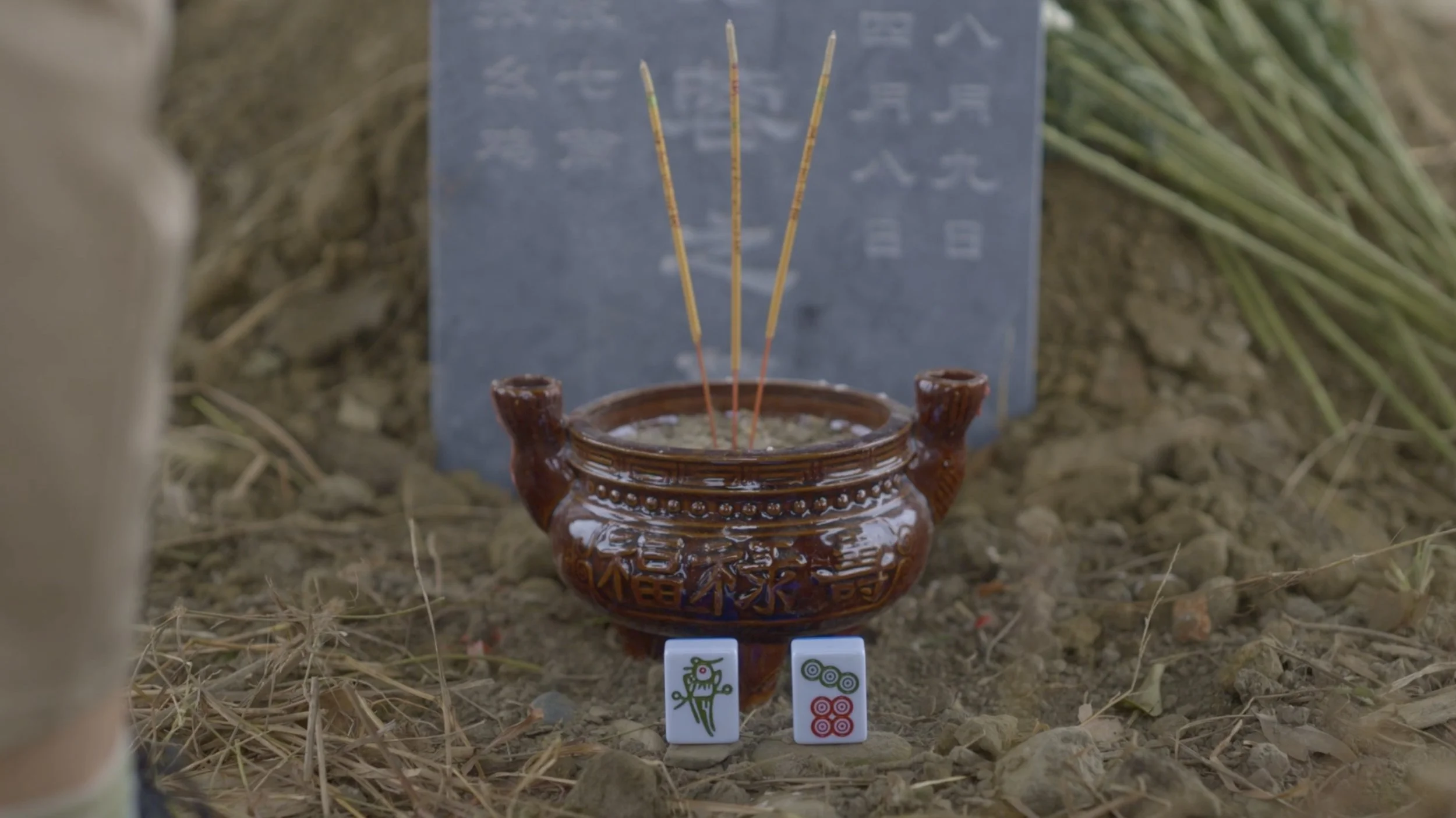 Ceremonial clay pot with incense sticks on the ground, with two Mahjong tiles in front, and a stone marker with inscriptions behind.