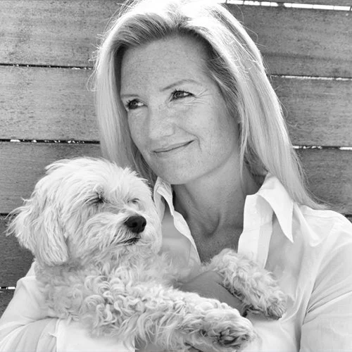 Marieke with long blonde hair smiling and holding a small fluffy dog in front of a wooden fence.