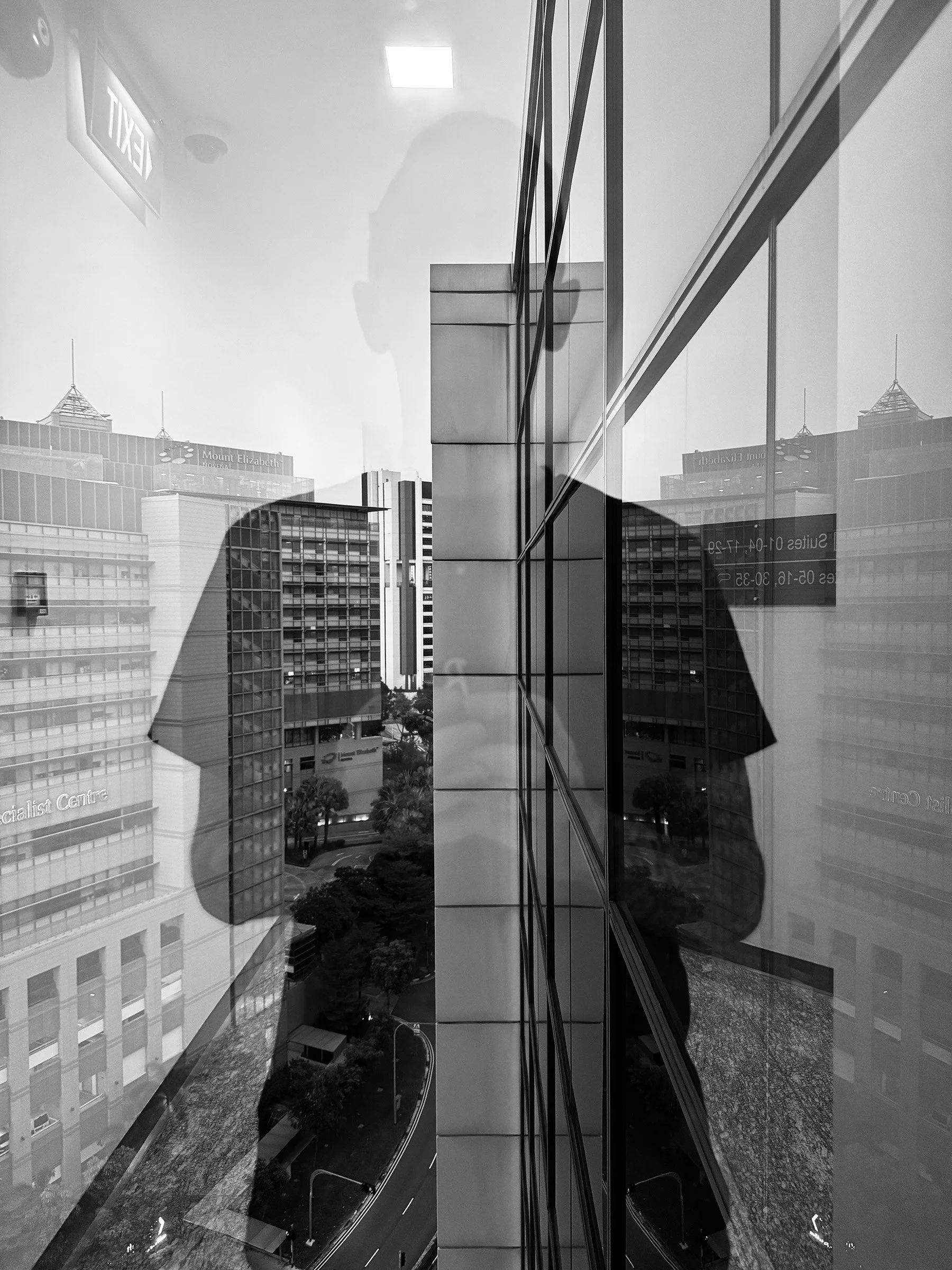 Black and white photo of a cityscape reflected in the glass windows of a building, with multiple tall office buildings and a street below.
