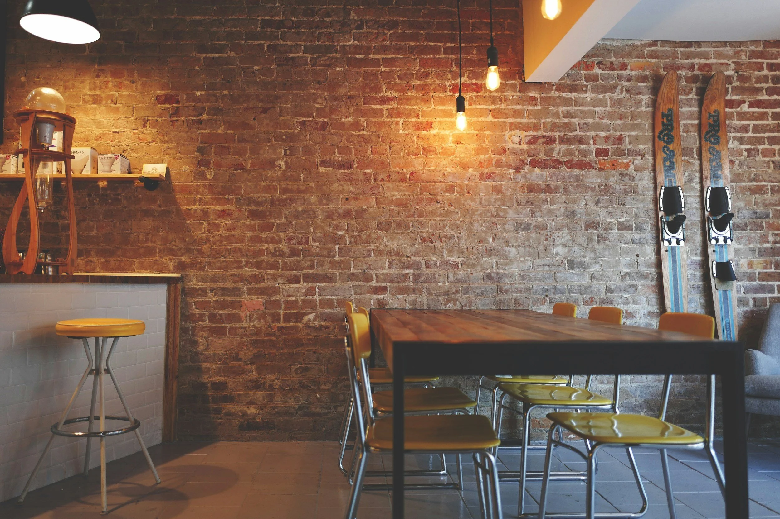 Interior of a cozy coffee shop with exposed brick walls, a wooden table surrounded by yellow chairs, vintage ceiling Edison bulbs, and a pair of vintage skis leaning against the brick wall.