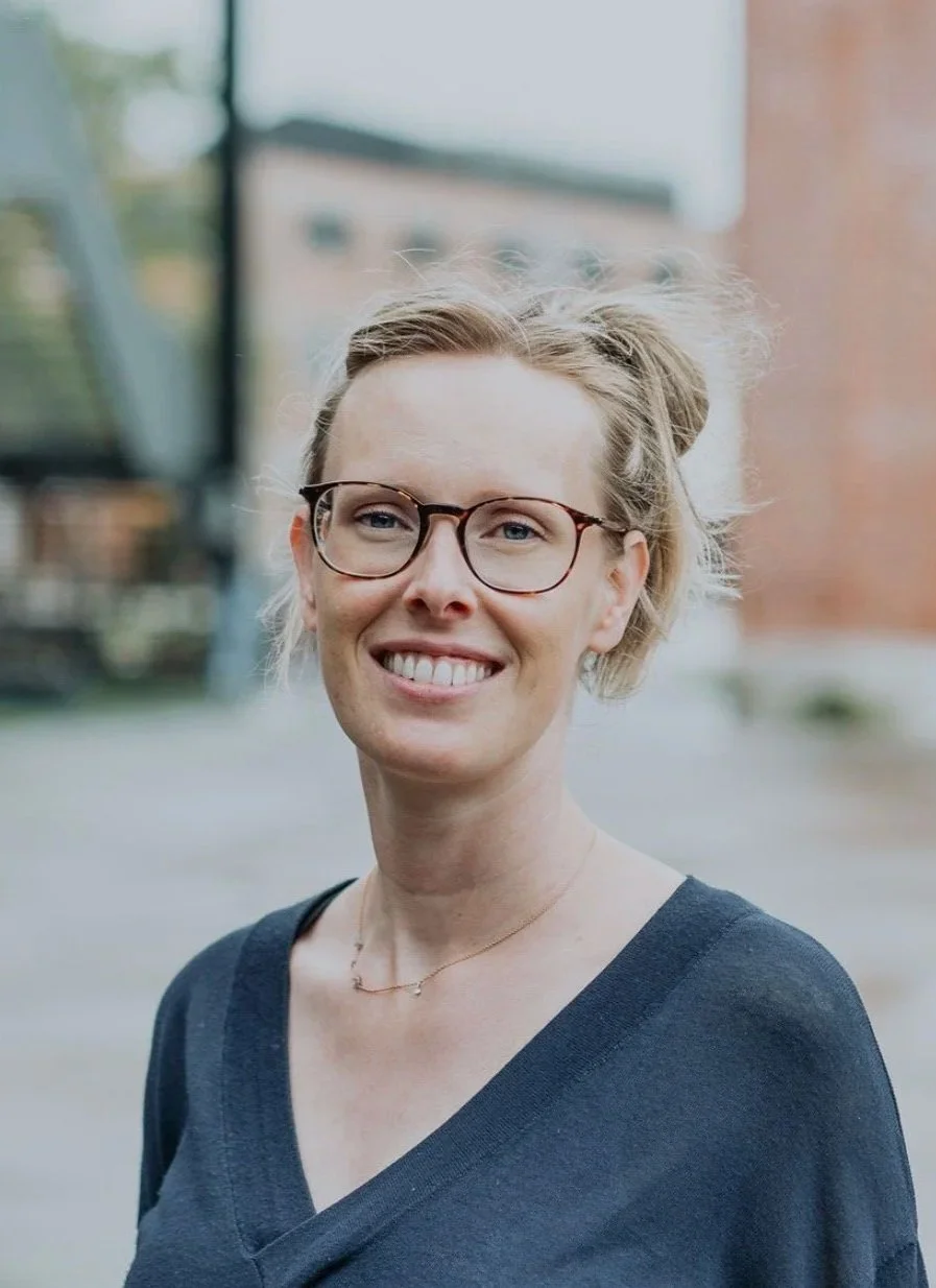 A smiling woman with glasses and wavy blonde hair in a bun, wearing a dark V-neck top and a delicate necklace, standing outdoors with a blurred background of a brick building and trees.