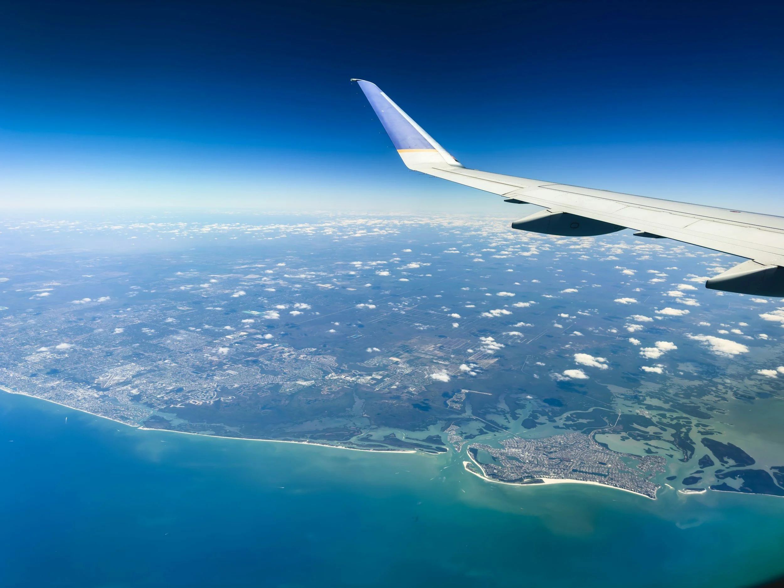 View from an airplane window showing the airplane wing and the coastline below, with blue sky and scattered clouds.