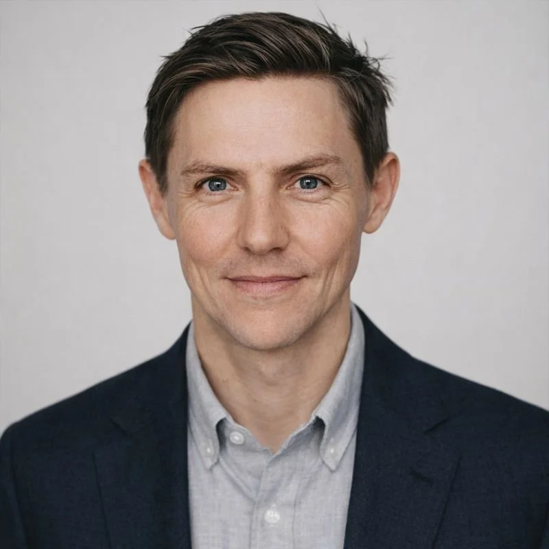 Headshot of a man with brown hair, blue eyes, wearing a dark blazer and light gray button-up shirt, against a plain background.