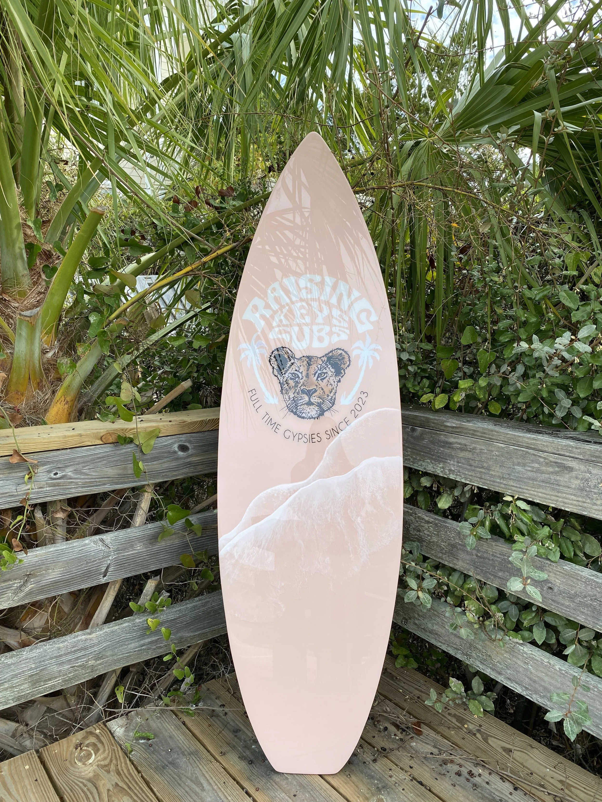 Pink surfboard with a lion head graphic and the words 'Tropical Vibes' resting against a wooden fence among green foliage.