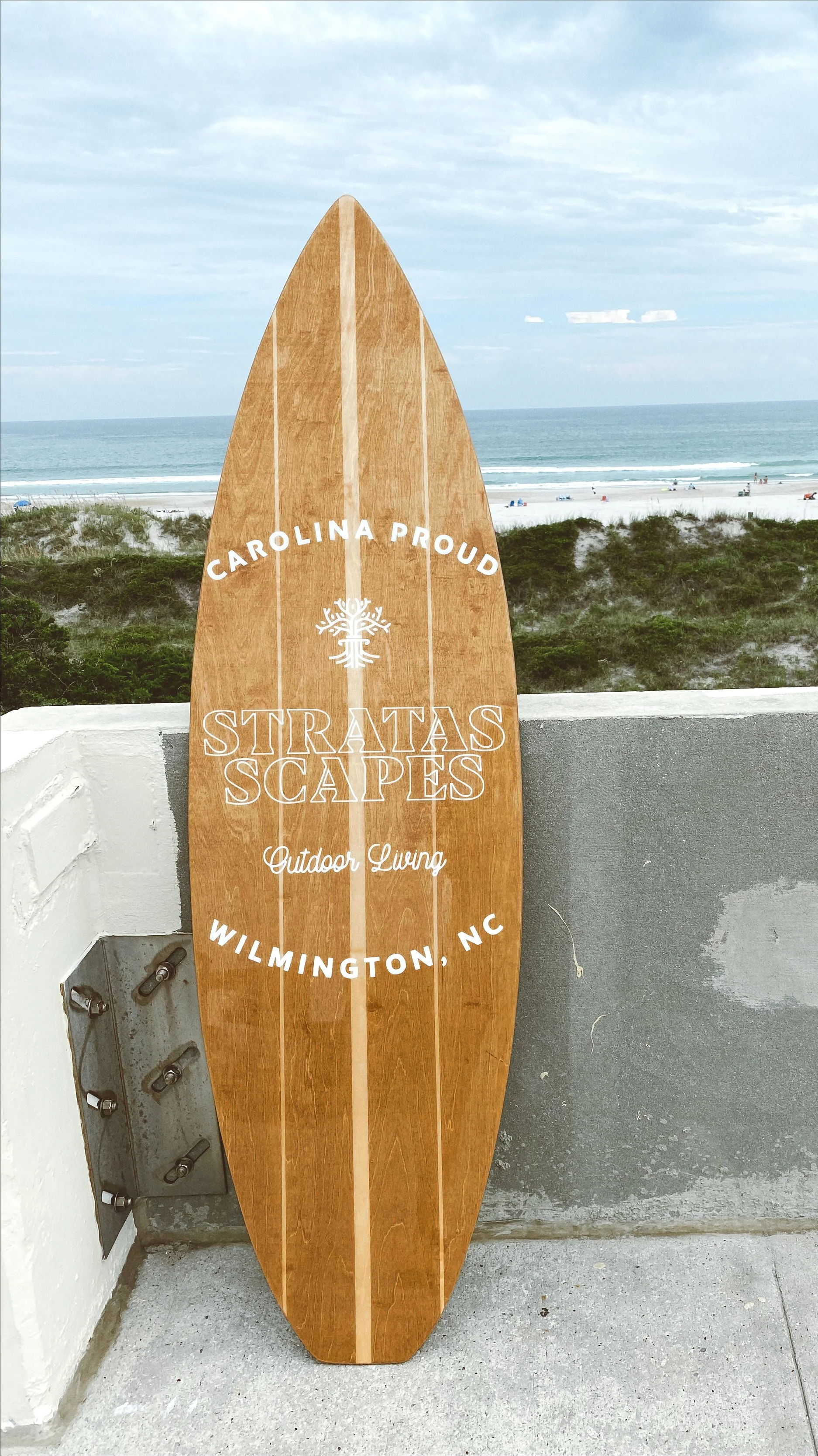 Wooden surfboard-shaped sign with text 'Carolina Proud', 'Stratas Scapes', 'Outdoor Living', and 'Wilmington, NC' against a beach background.