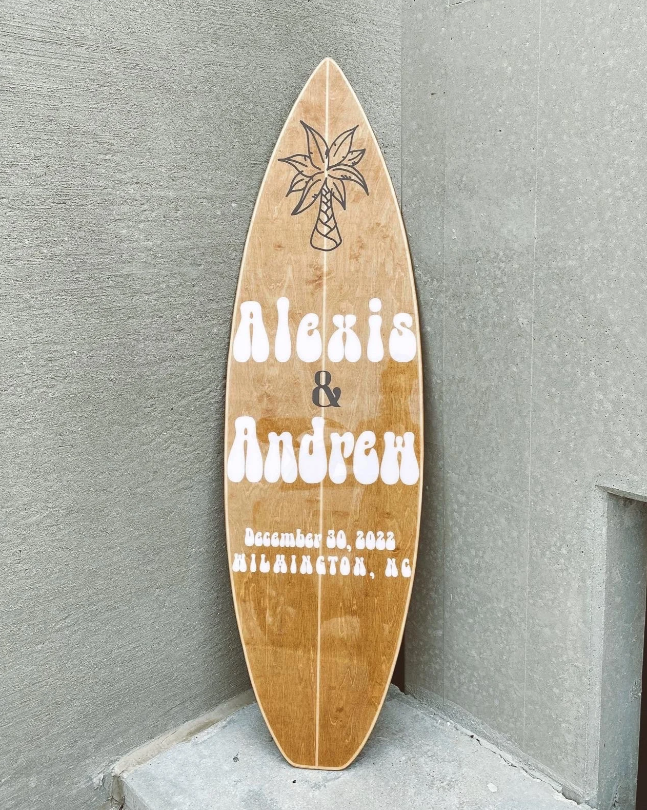 Wooden surfboard with white and black text, featuring a drawing of a palm tree at the top, used as a sign for a wedding event for Alexis & Andrew on December 30, 2022, in Wilmington, North Carolina.
