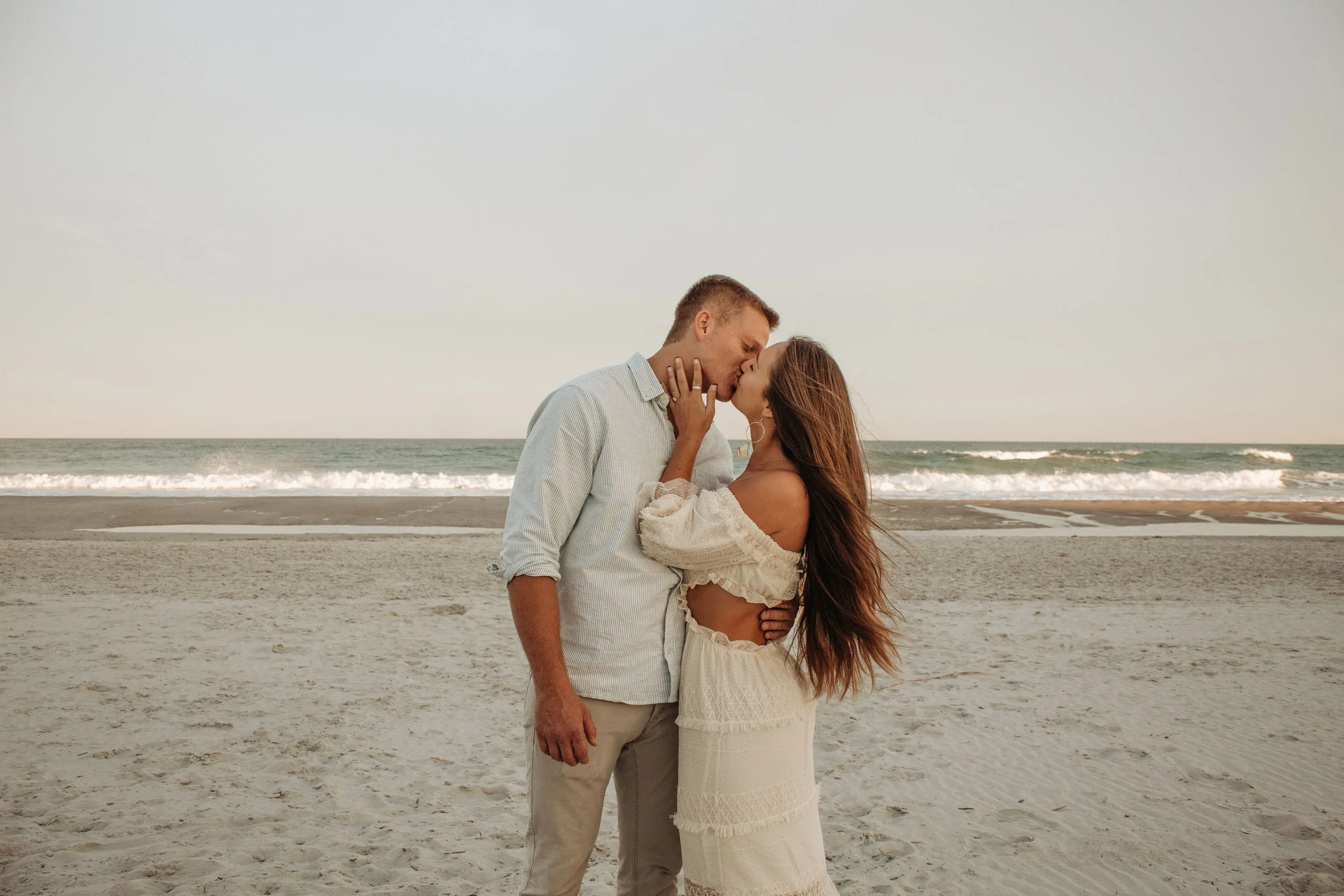 A couple sharing a kiss on the beach at sunset, with the ocean in the background.